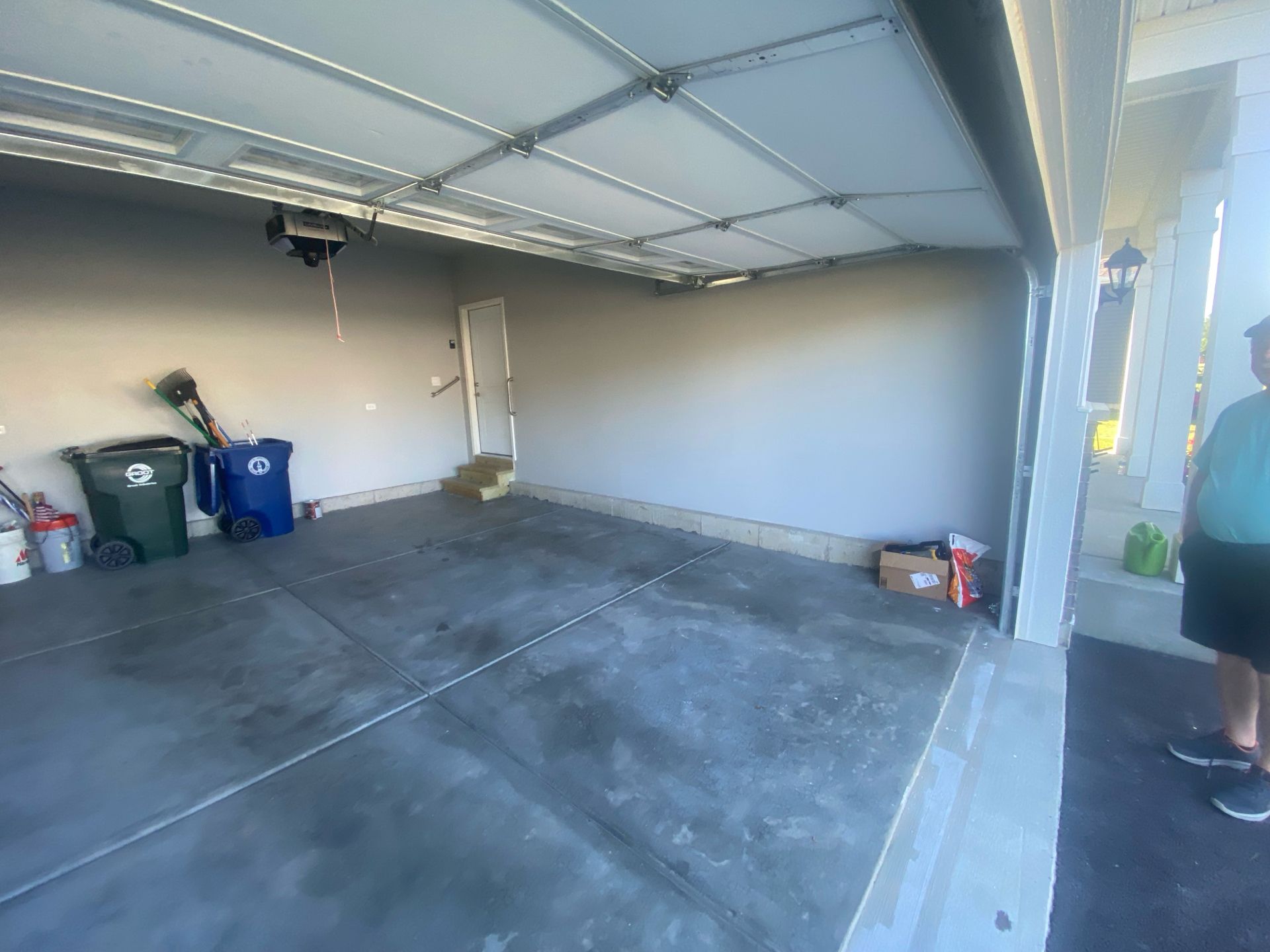 An empty, two-car garage with a concrete floor, two bins in the back left, and a person standing on the right.