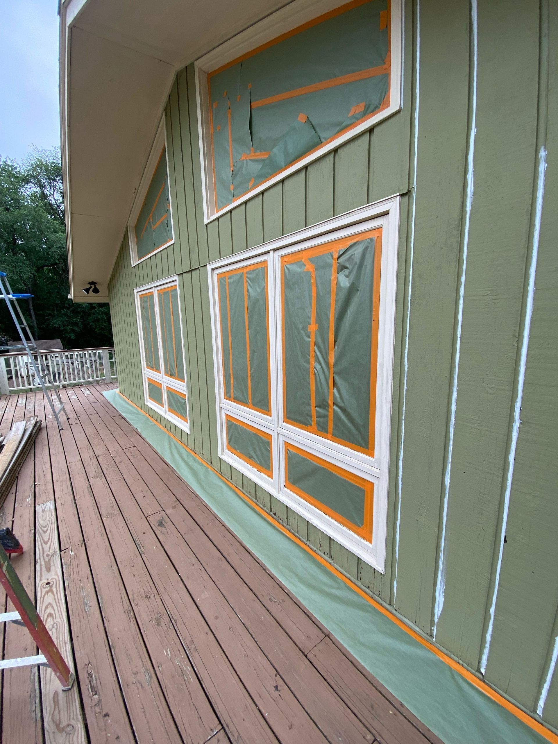 Exterior wall under renovation, featuring olive-green siding with white vertical seams, and windows masked with tape.