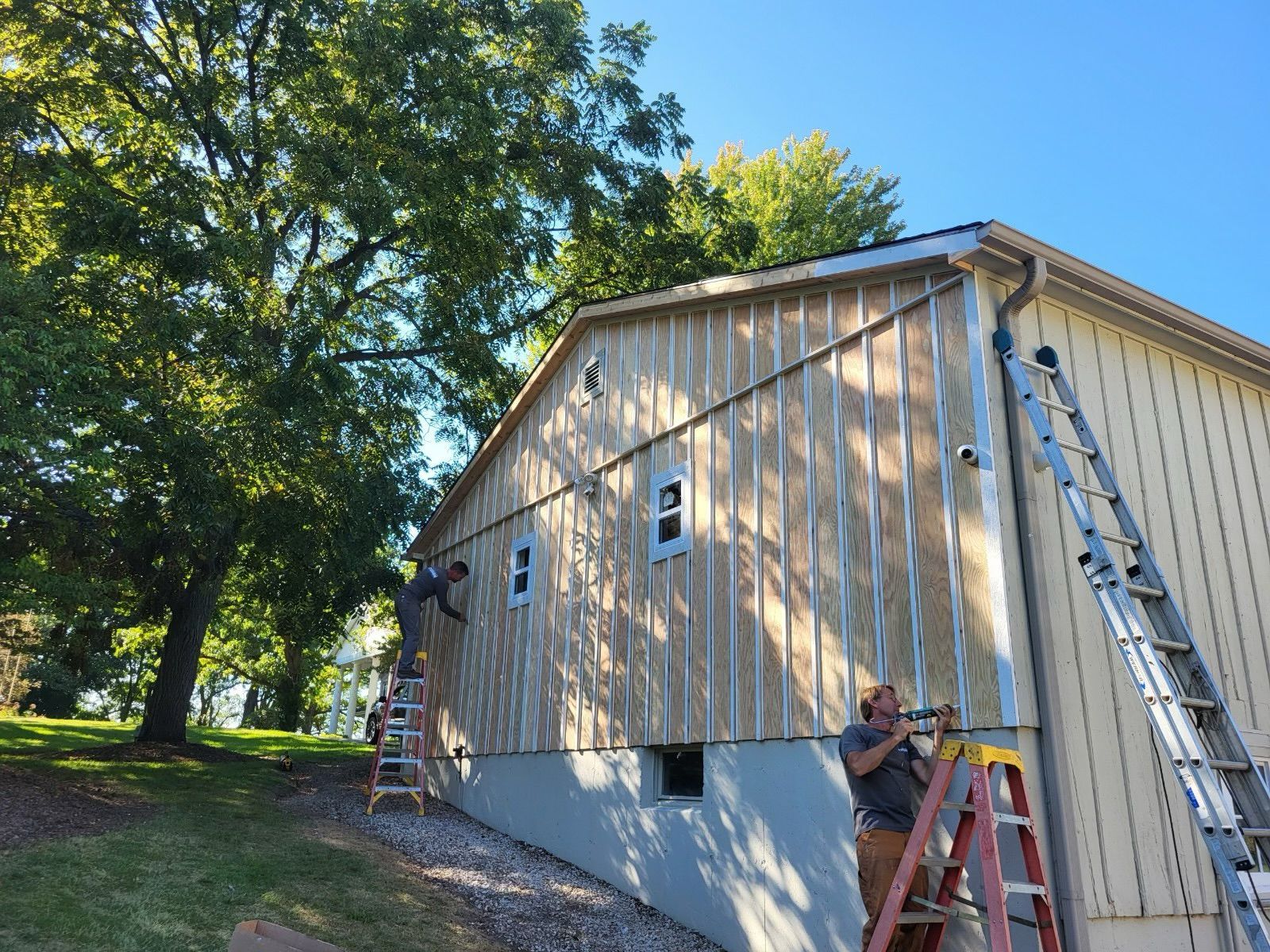Two people work on a ladder and scaffolding to install vertical siding on the light-colored exterior wall of a house.