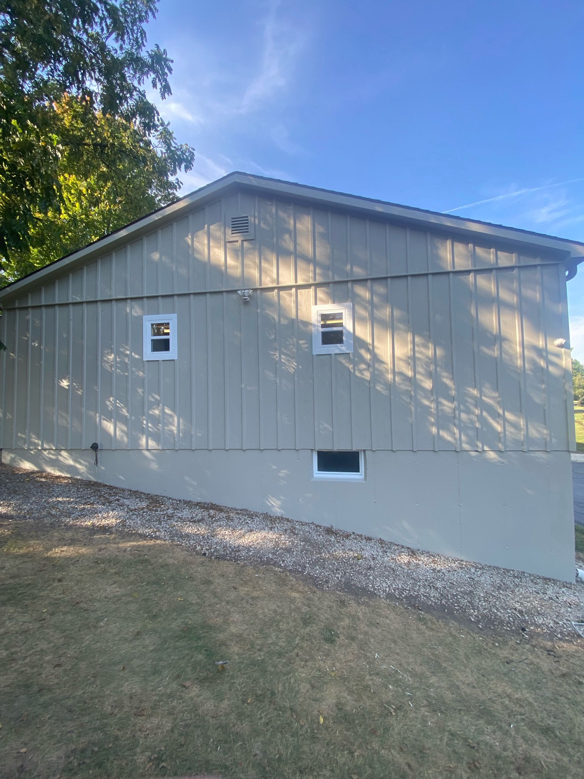 Beige side of a house with two upper windows, a basement window, and a gravel slope against a foundation under blue sky.