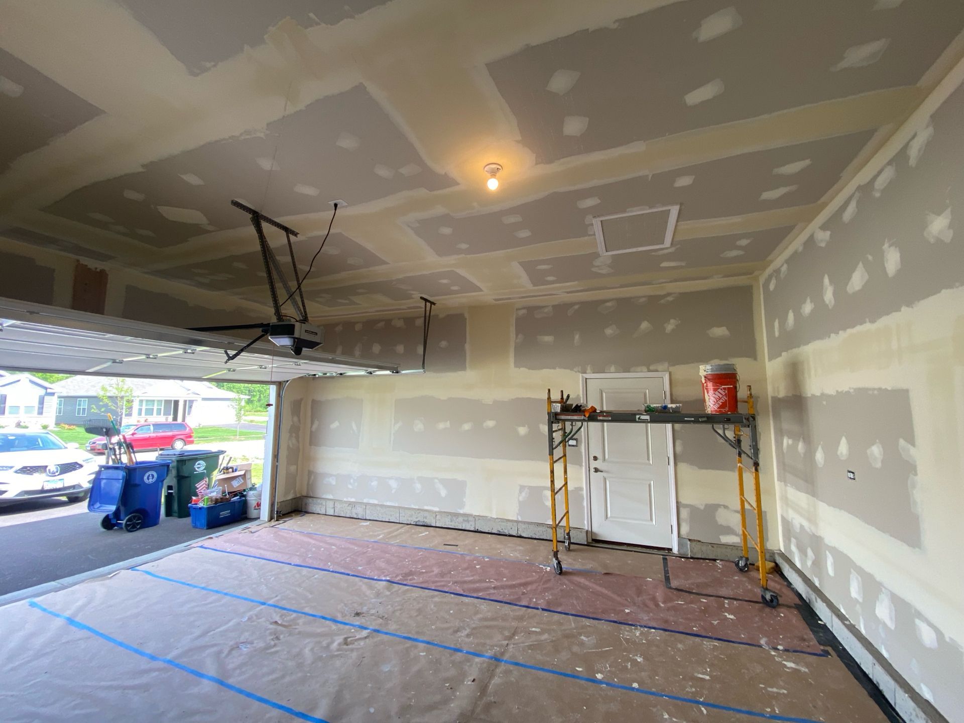 Interior of a residential garage during drywall installation, featuring unfinished taped seams and a mobile scaffold.