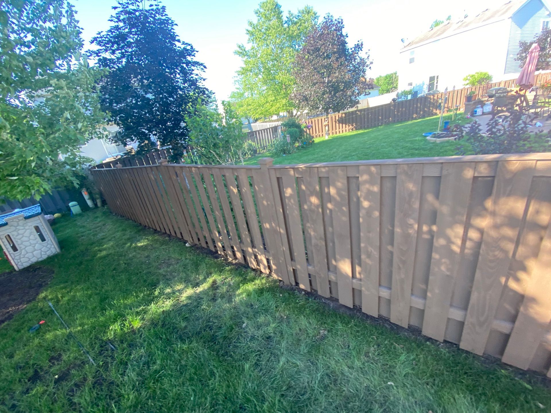 A brown wooden fence runs across a sunny, green backyard with trees and a small plastic playhouse on the left.