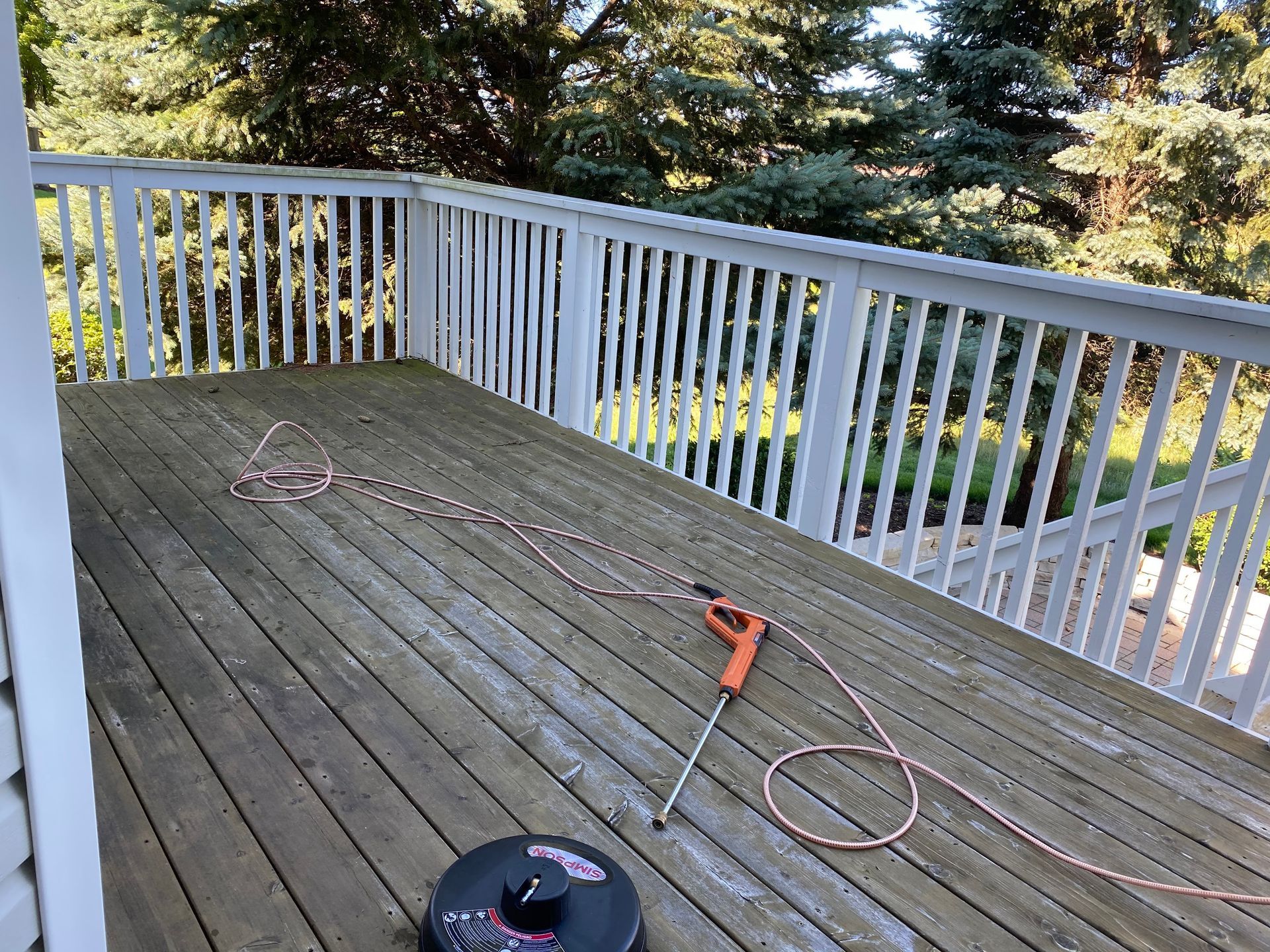 A power washing wand and circular surface cleaner sit on a weathered wooden deck with a white railing.