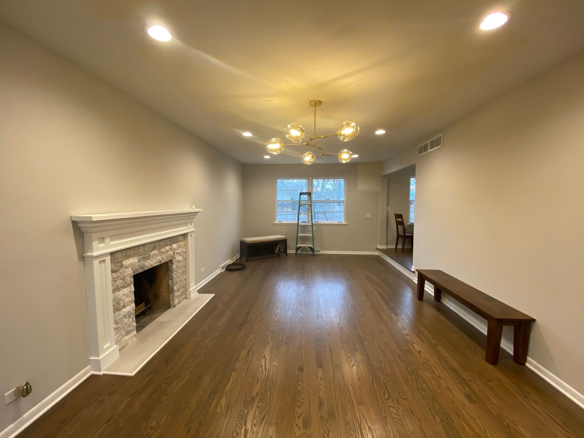 A long, empty room with light gray walls, dark wood floors, a stone fireplace, and a hanging chandelier.