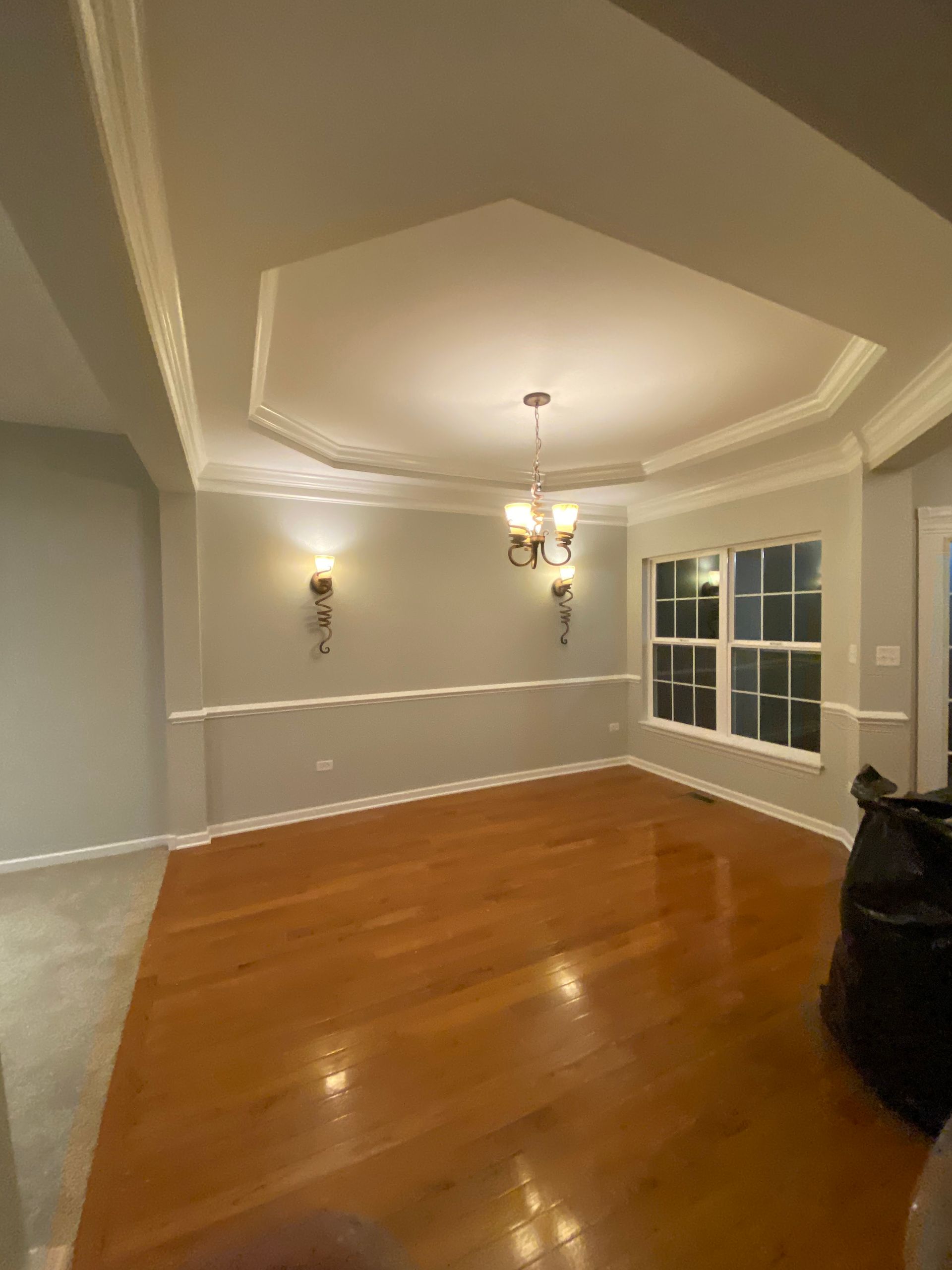 An empty dining room with hardwood floors, a tray ceiling with a chandelier, wall sconces, and gray walls.