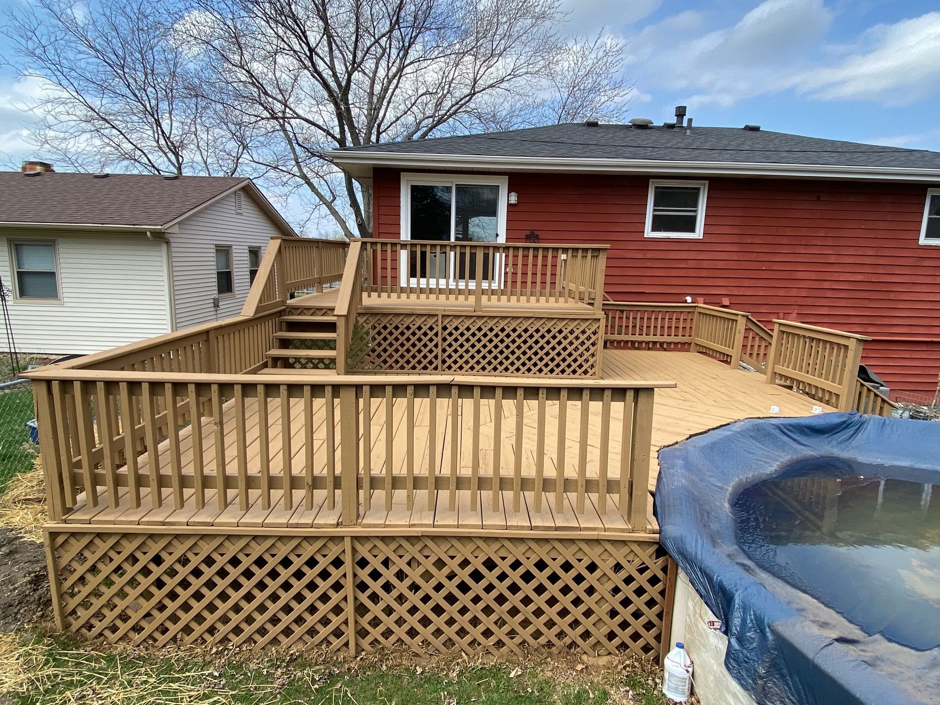 A multi-level tan wooden deck with lattice skirting sits against a red house next to a pool covered with a blue tarp.
