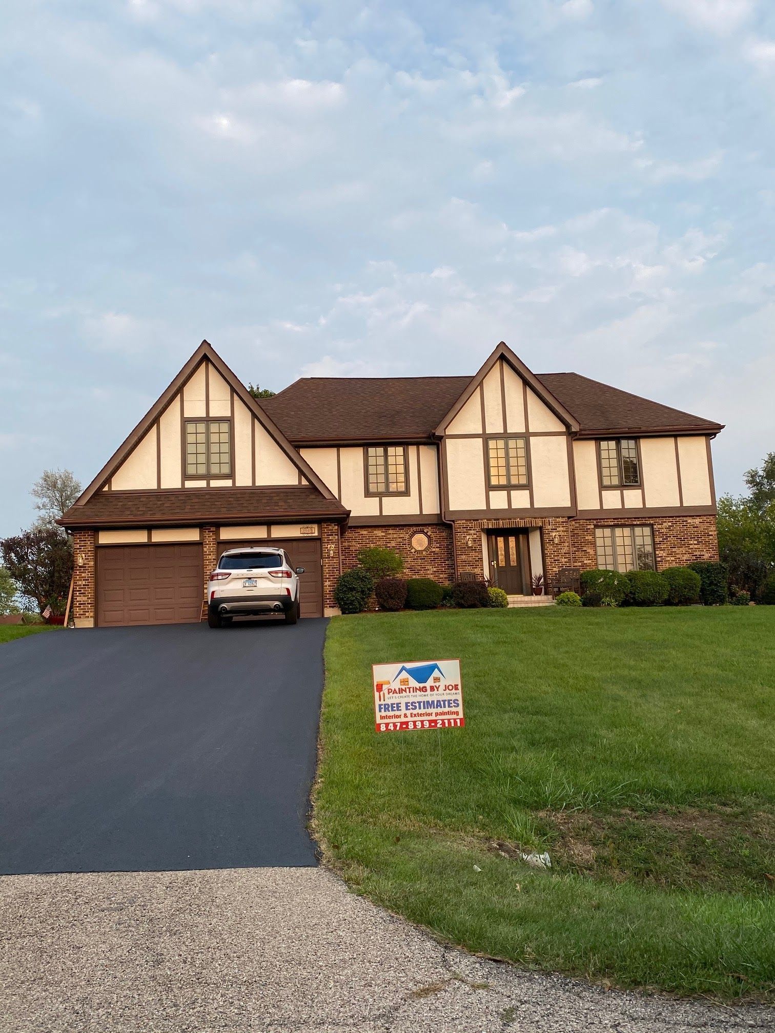 A two-story Tudor-style home with light stucco, dark brown timber accents, a brown roof, and a paved driveway with a car.