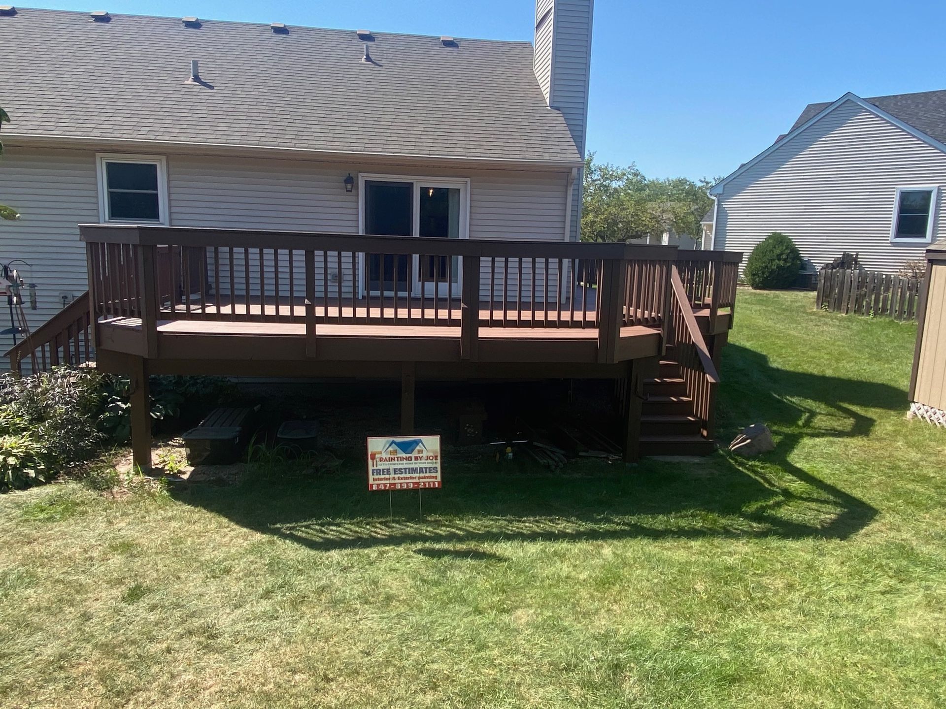 A brown wooden deck attached to the back of a light-colored house with a yard on a sunny day.