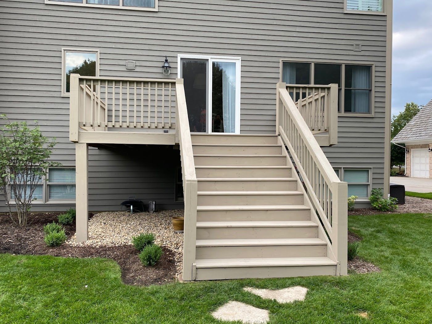 A tan wooden deck with stairs leading to a sliding glass door on the back of a house with grey horizontal siding.