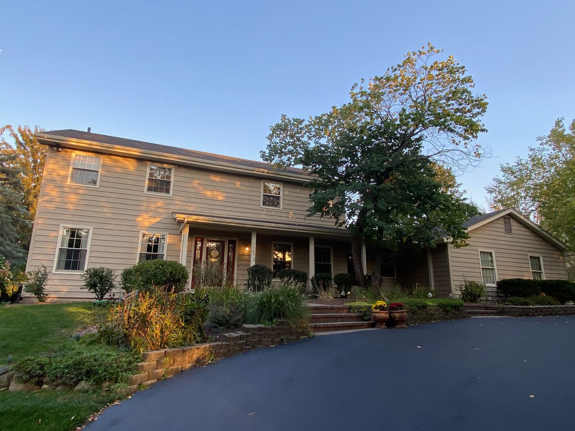 Two-story house with light tan brick, a front porch, and mature trees during a golden sunset, viewed from the driveway.