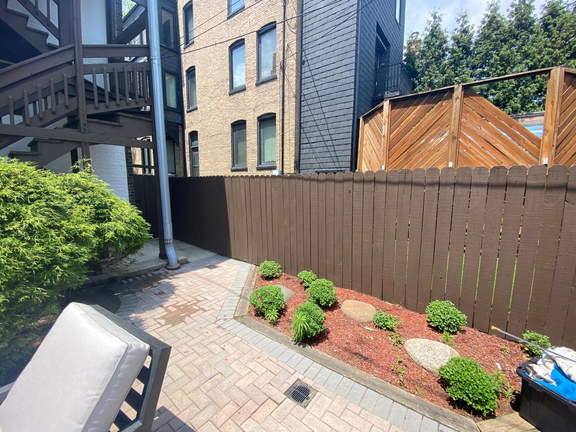 A brick patio area with a small garden bed featuring small green shrubs, wood chips, and a brown wooden privacy fence.