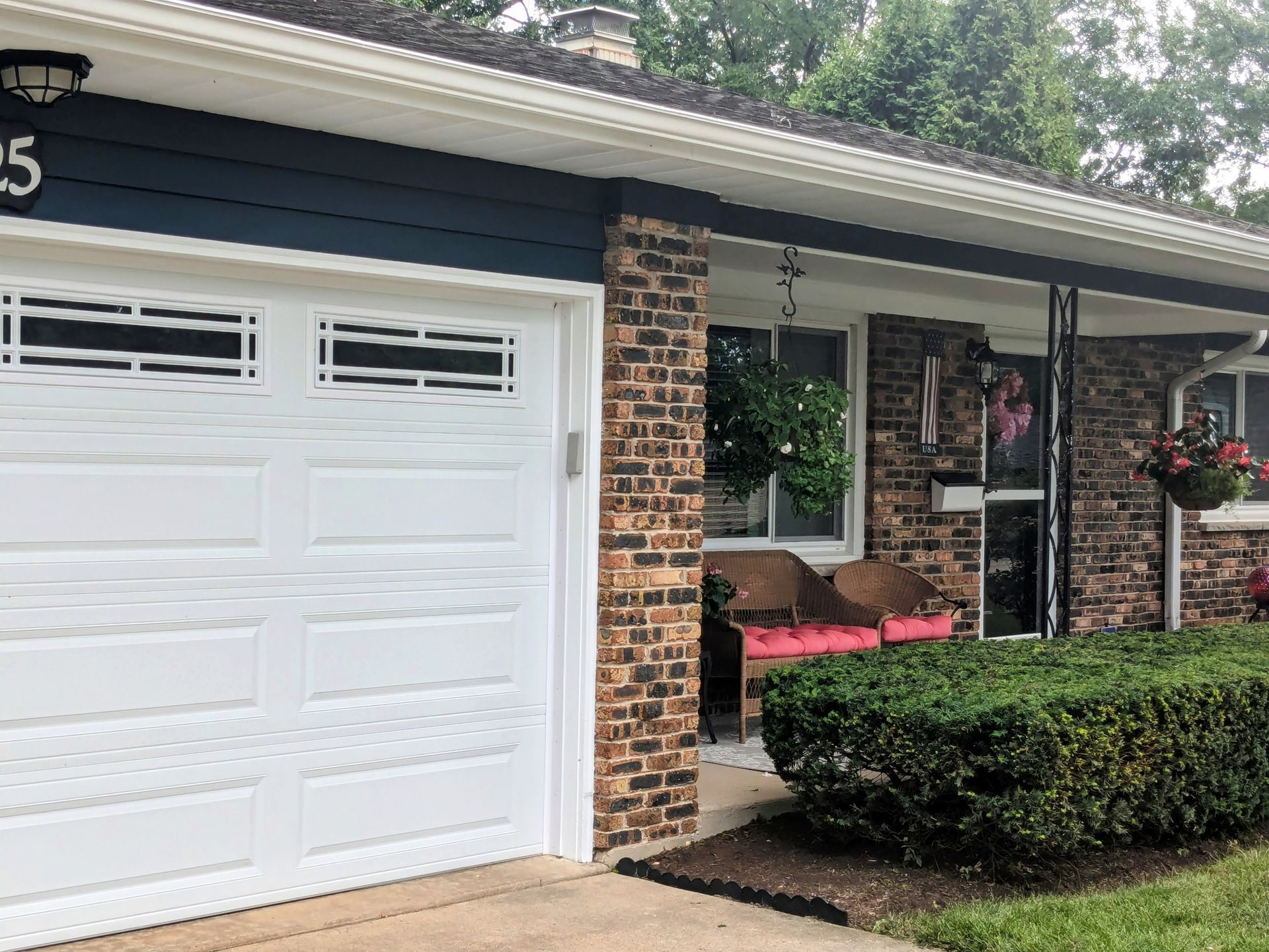 A front view of a house with a white garage door, dark blue siding, and a brick exterior with a small porch and hedges.