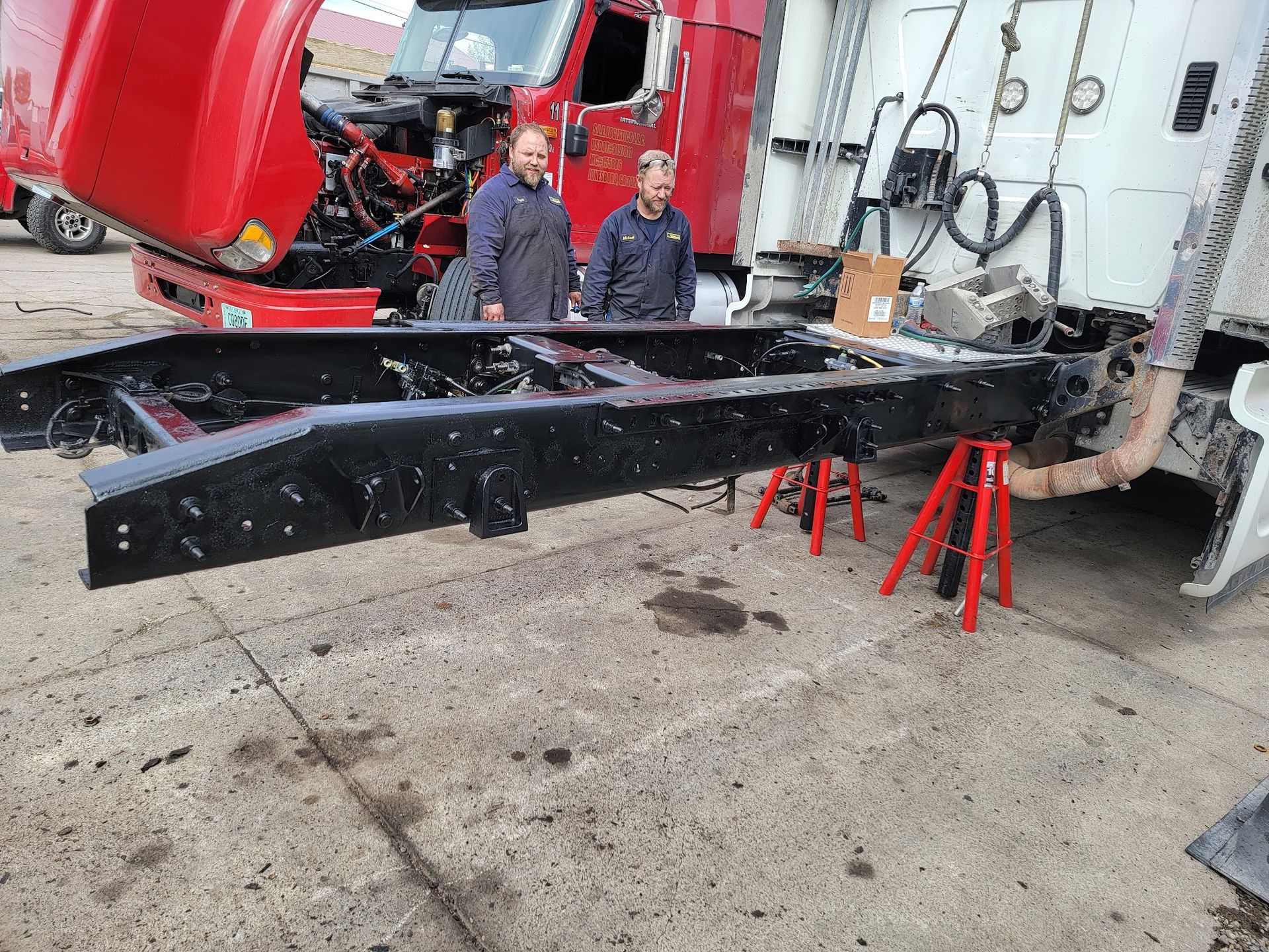 Two men in work clothes stand near a truck frame supported on jack stands.