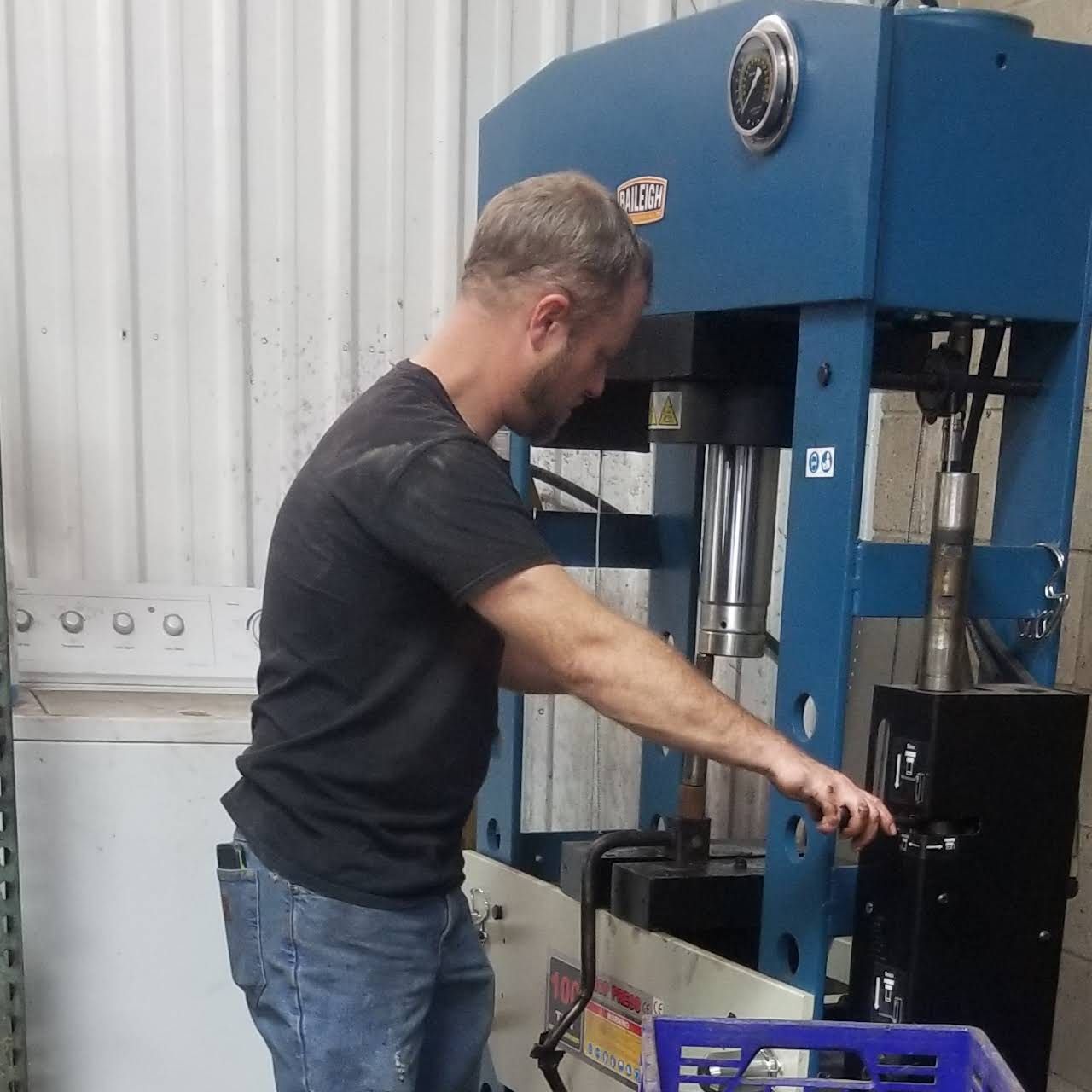 Man operating a large blue industrial press in a workshop, wearing a black shirt and jeans.
