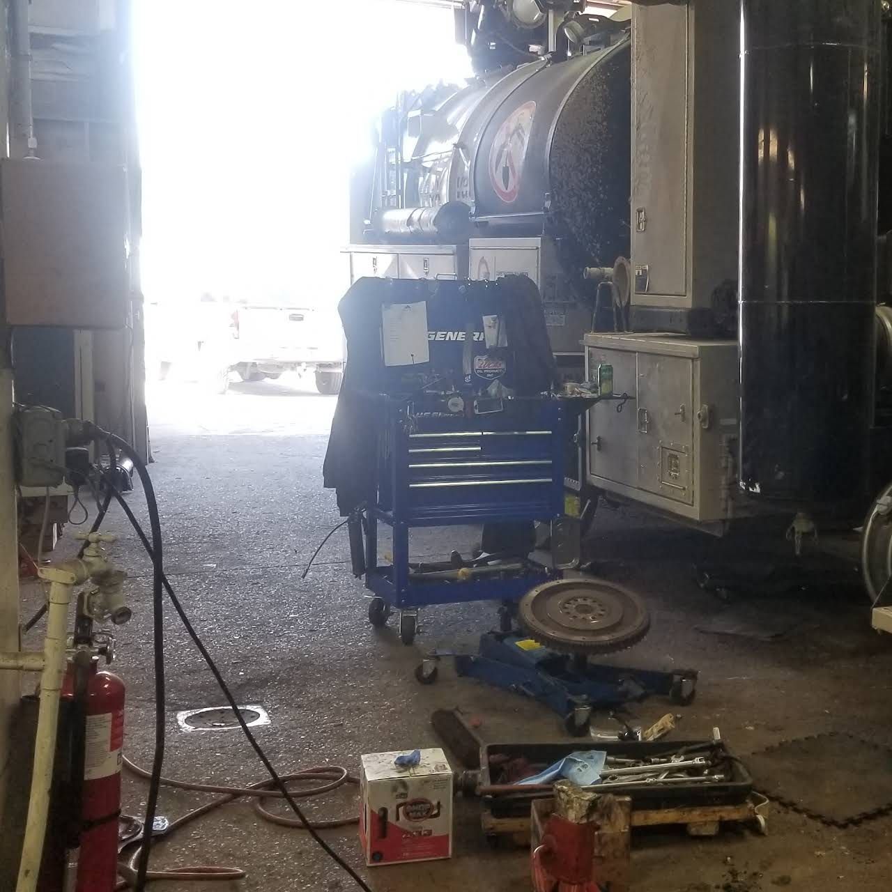 Mechanic's bay with a truck undergoing repair. Tools and equipment are scattered around. Bright doorway.