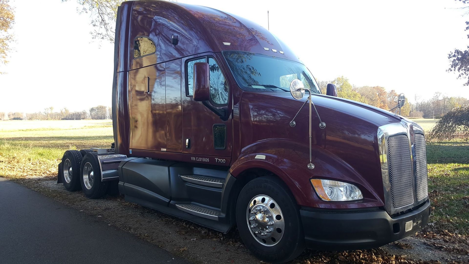Maroon semi-truck parked on the side of a road with a green field in the background.