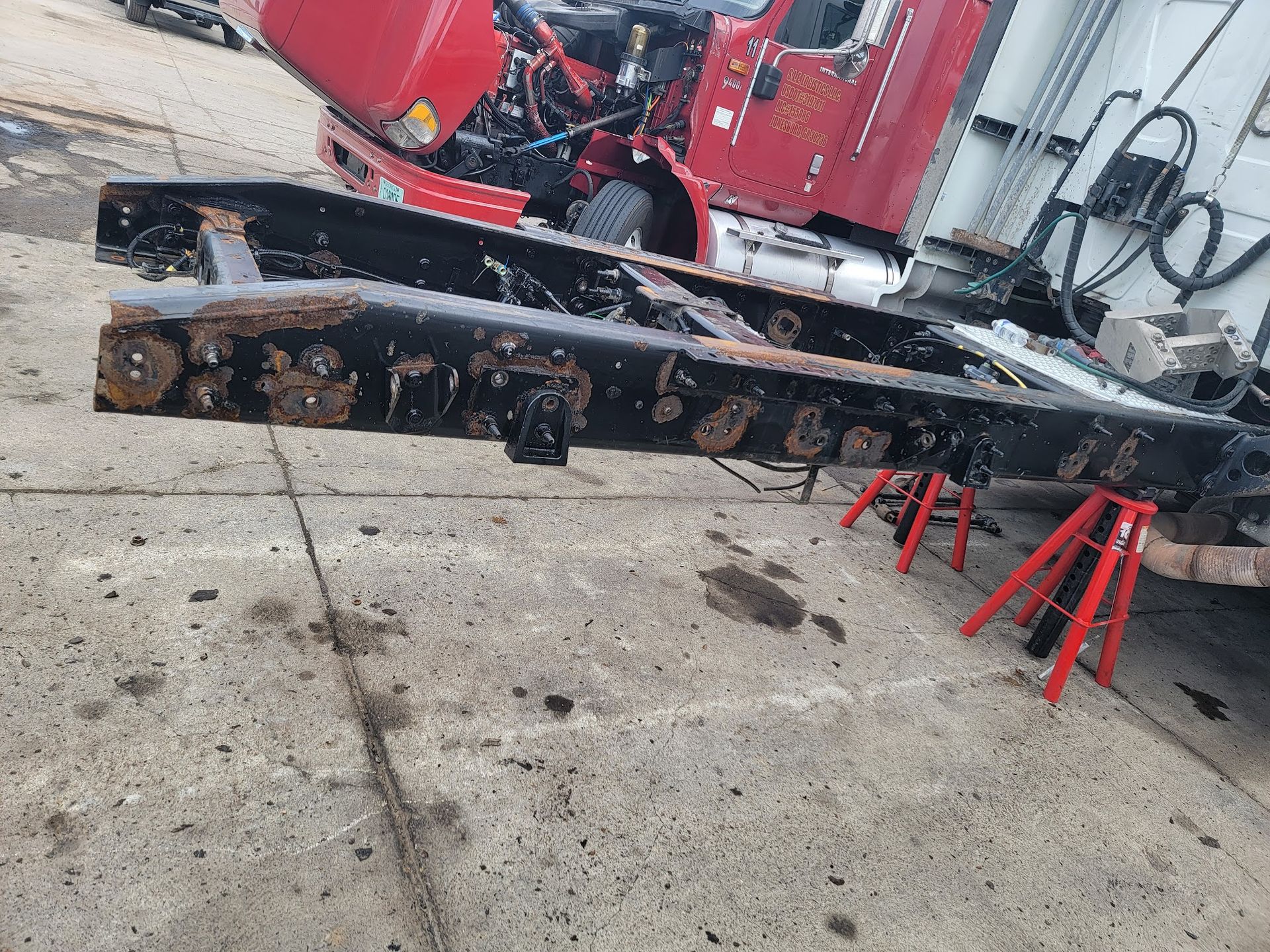 Black truck frame on jack stands, near a red truck and concrete surface.