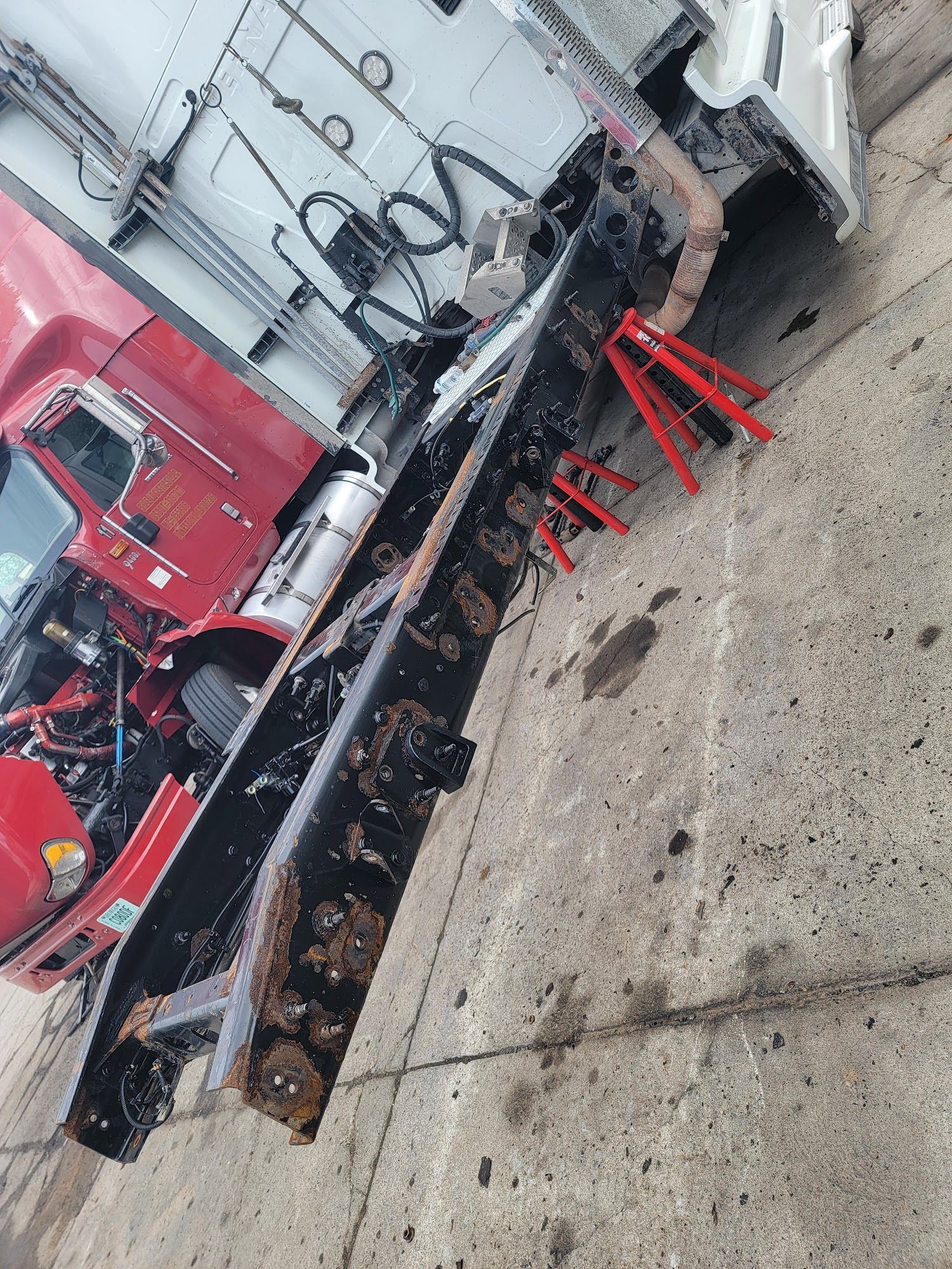 Red semi-truck frame with a detached, rusted section on concrete, supported by jack stands.