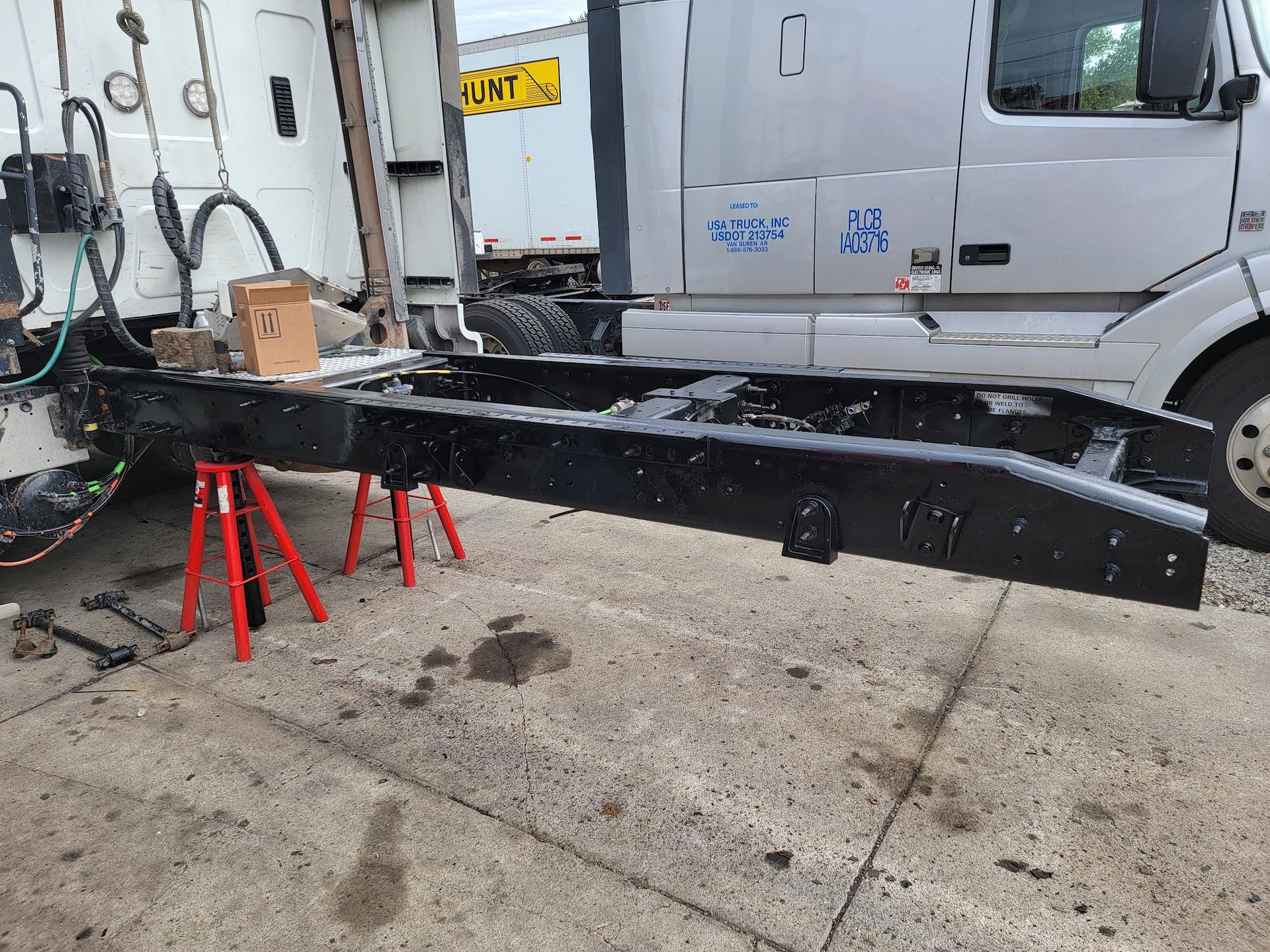 A black semi-truck frame is raised on red jack stands near a white truck.