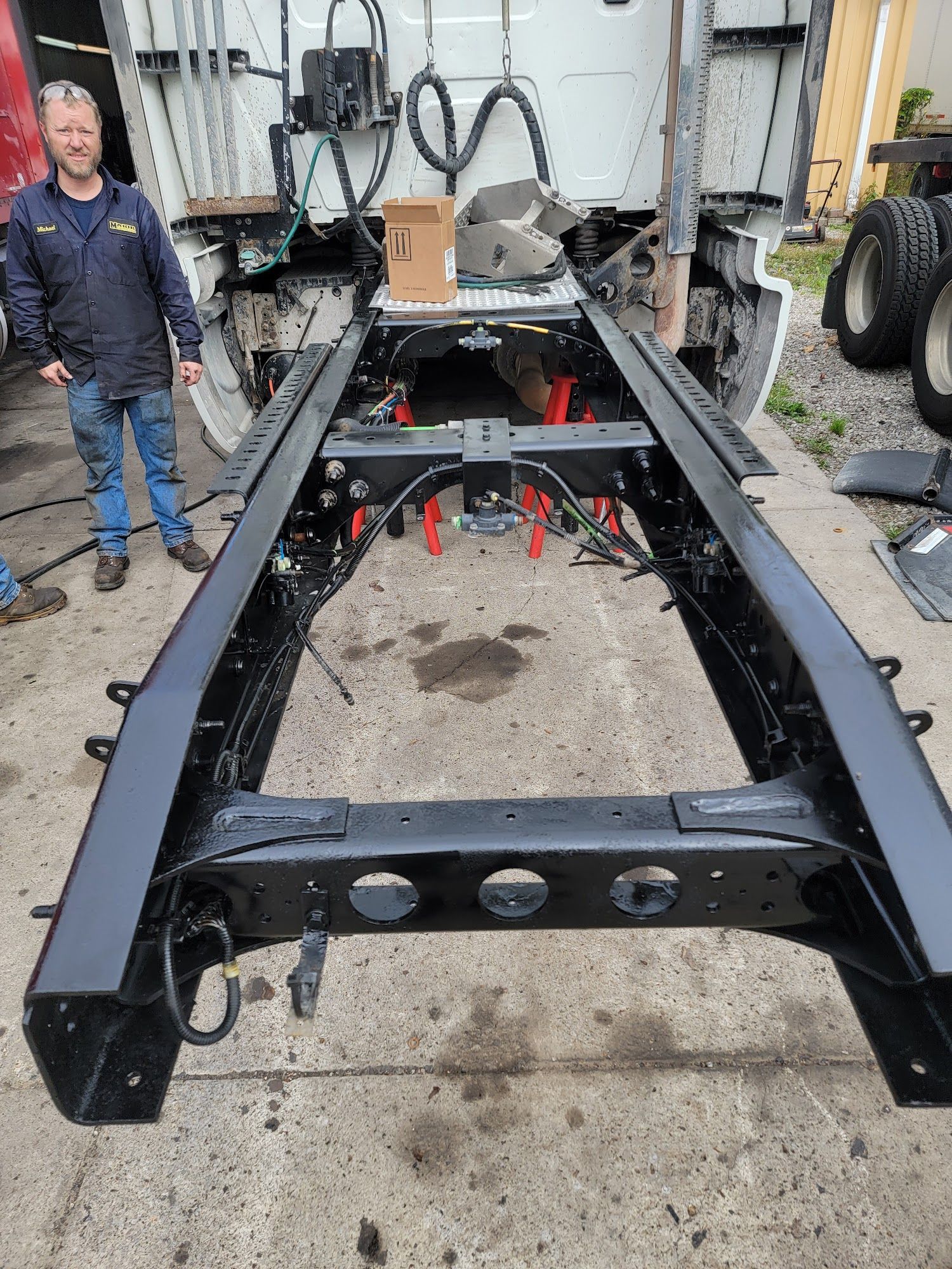 Man standing near a black truck frame on a concrete surface, red supports visible.