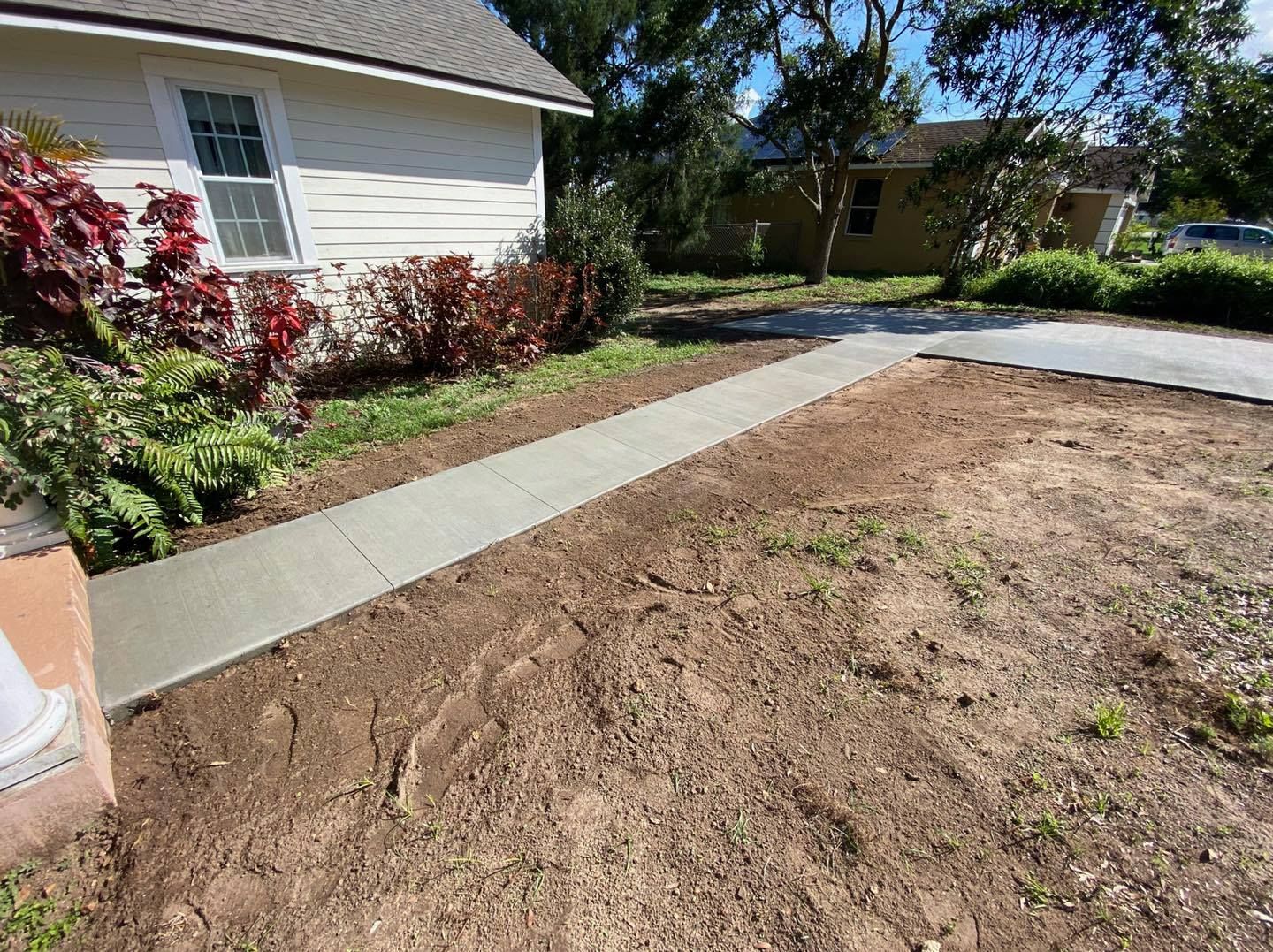 Newly poured concrete sidewalk leading to a house. Brown earth and greenery surround.