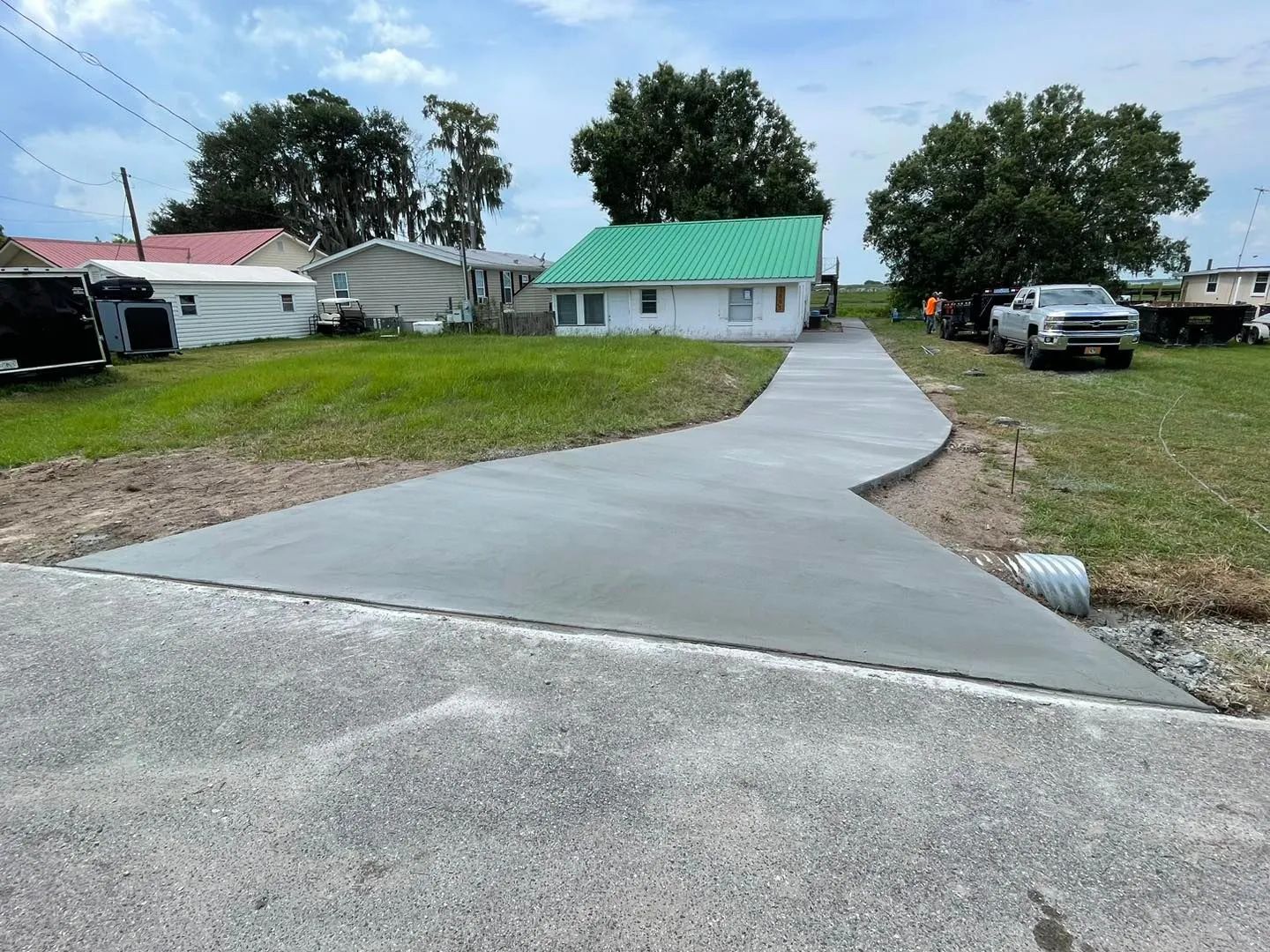 Newly poured concrete driveway leading to a white house with a green roof, on a grassy lot.