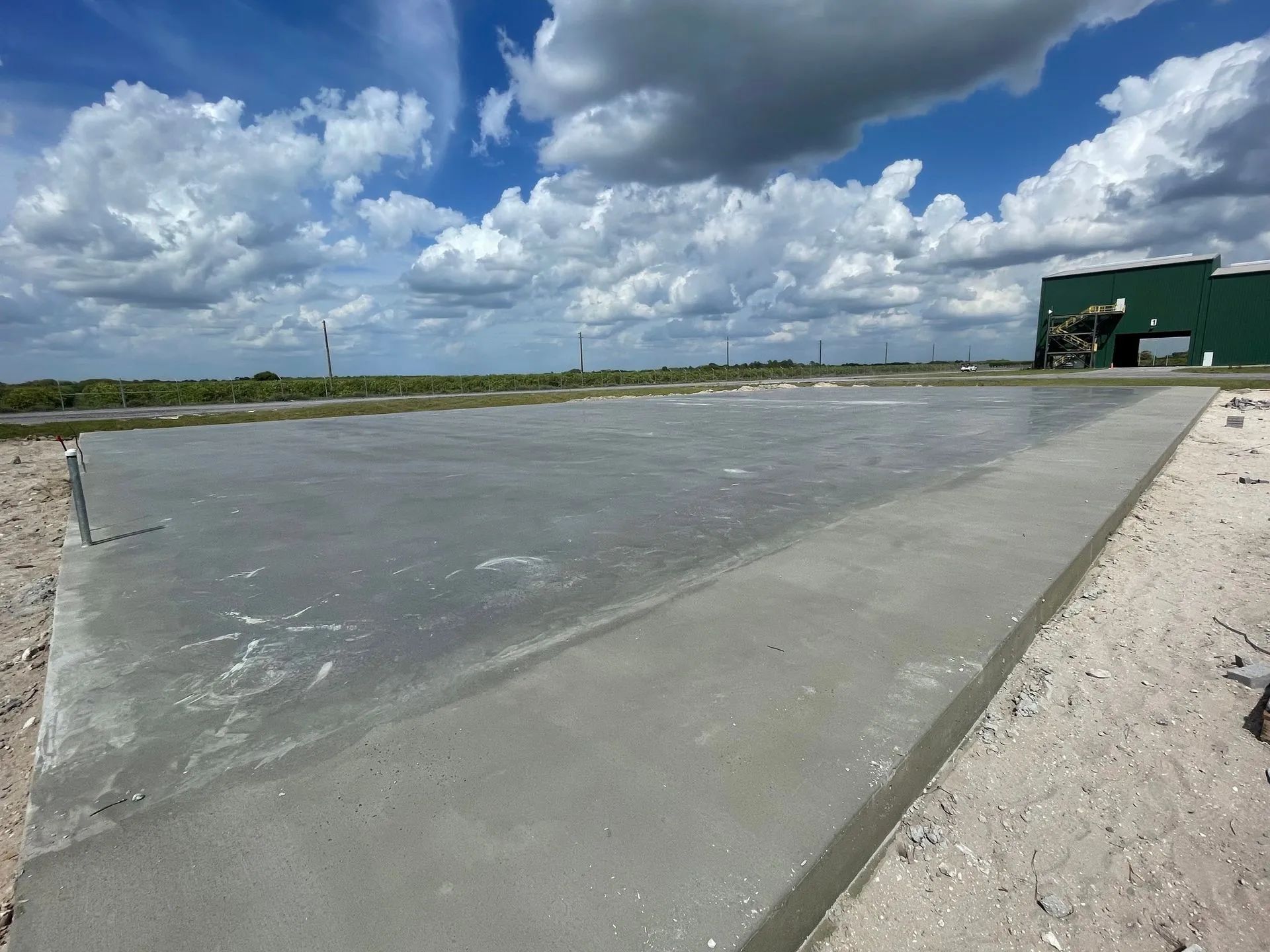 Newly poured concrete pad with a green building in the distance under a cloudy sky.
