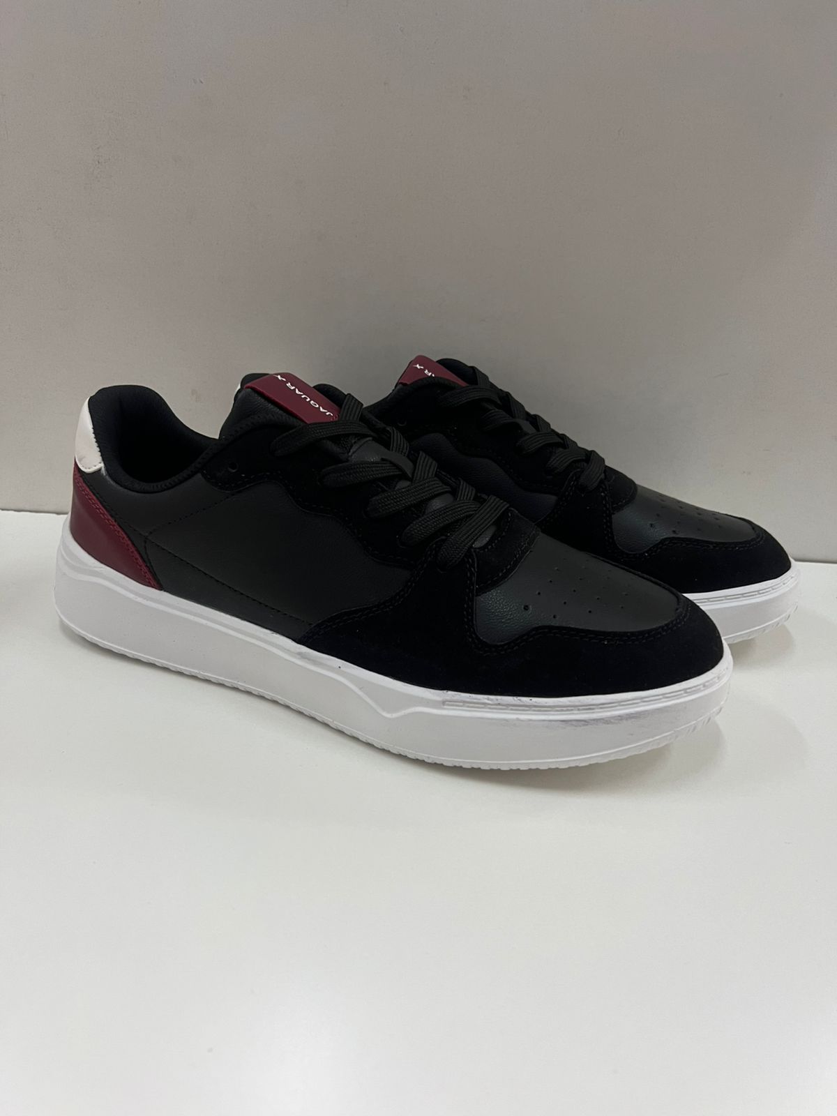 Zapatillas negras con suela blanca y detalles en color burdeos, sobre superficie blanca.