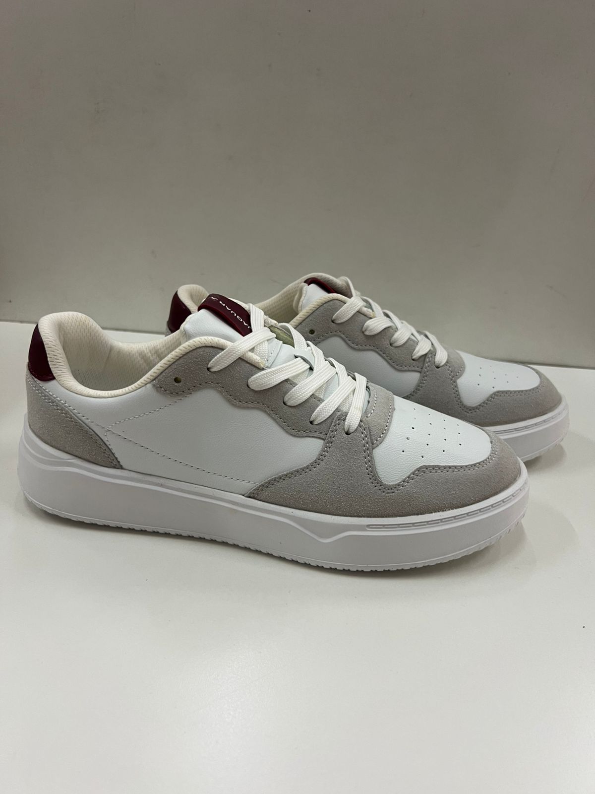 Zapatillas blancas con detalles de gamuza gris y detalle de talón burdeos sobre una superficie blanca.
