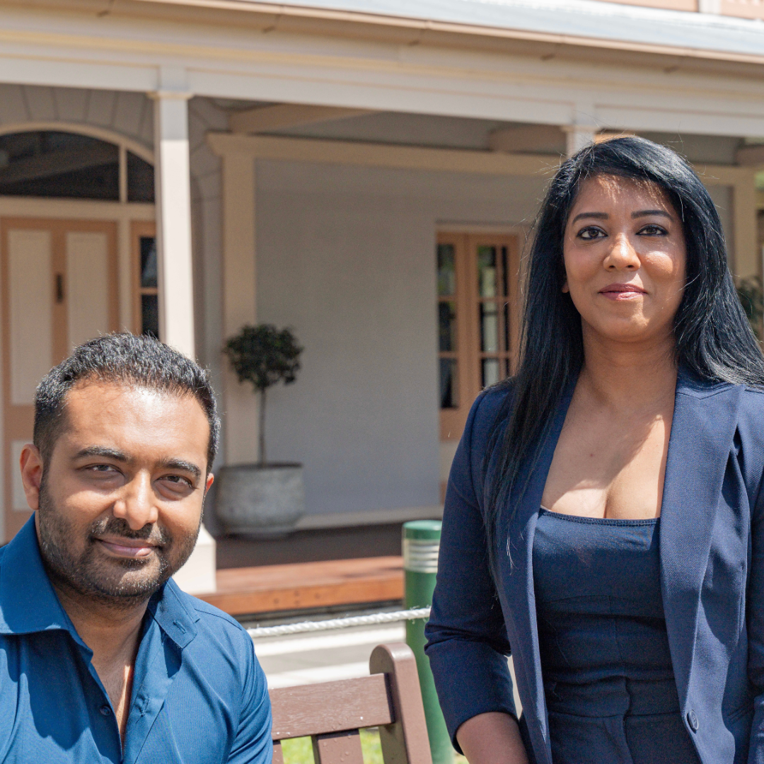 Man and woman smiling outdoors, in front of a building with a porch.