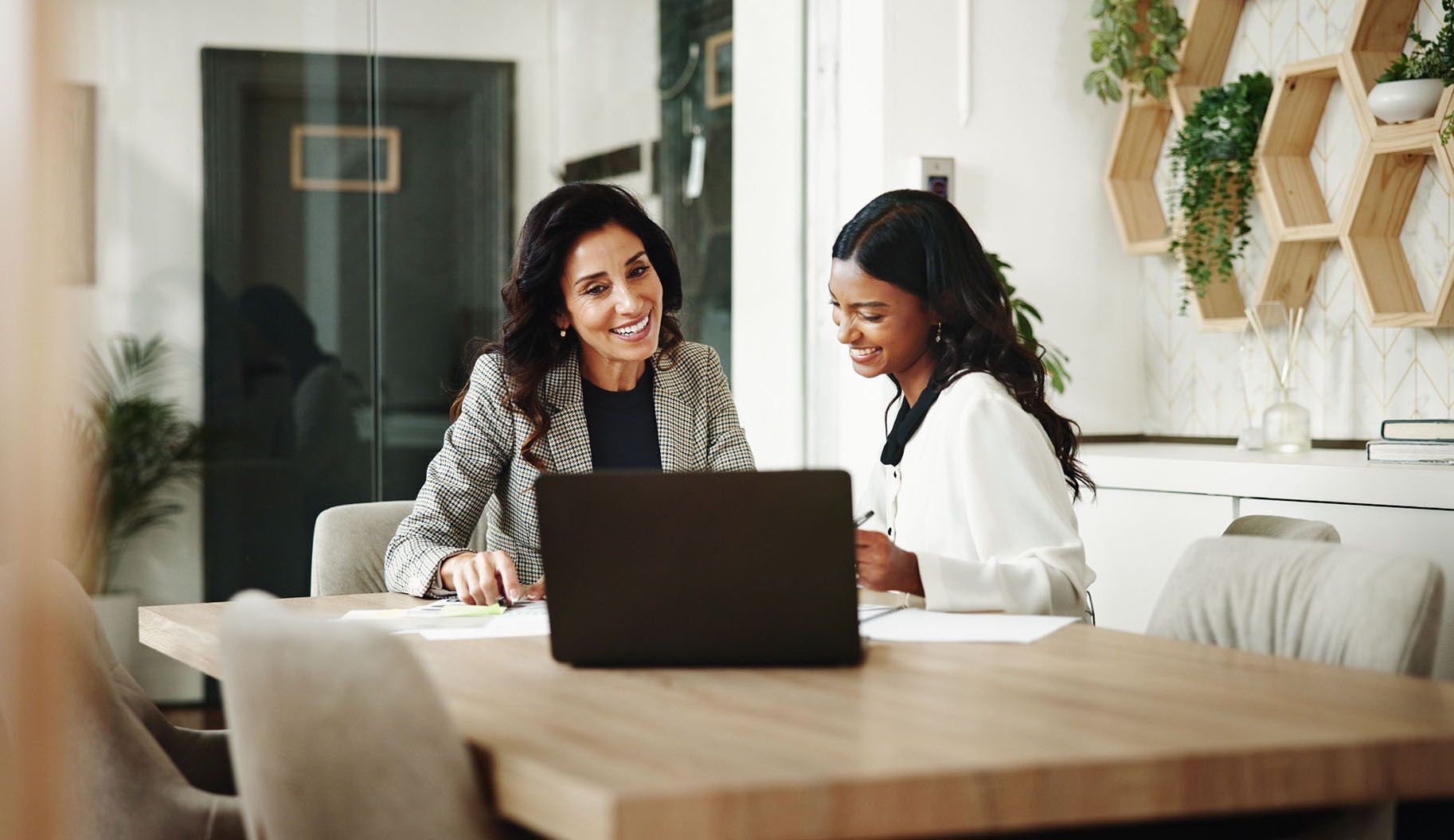 Two women smiling, working together at a table with a laptop and papers in a modern office.