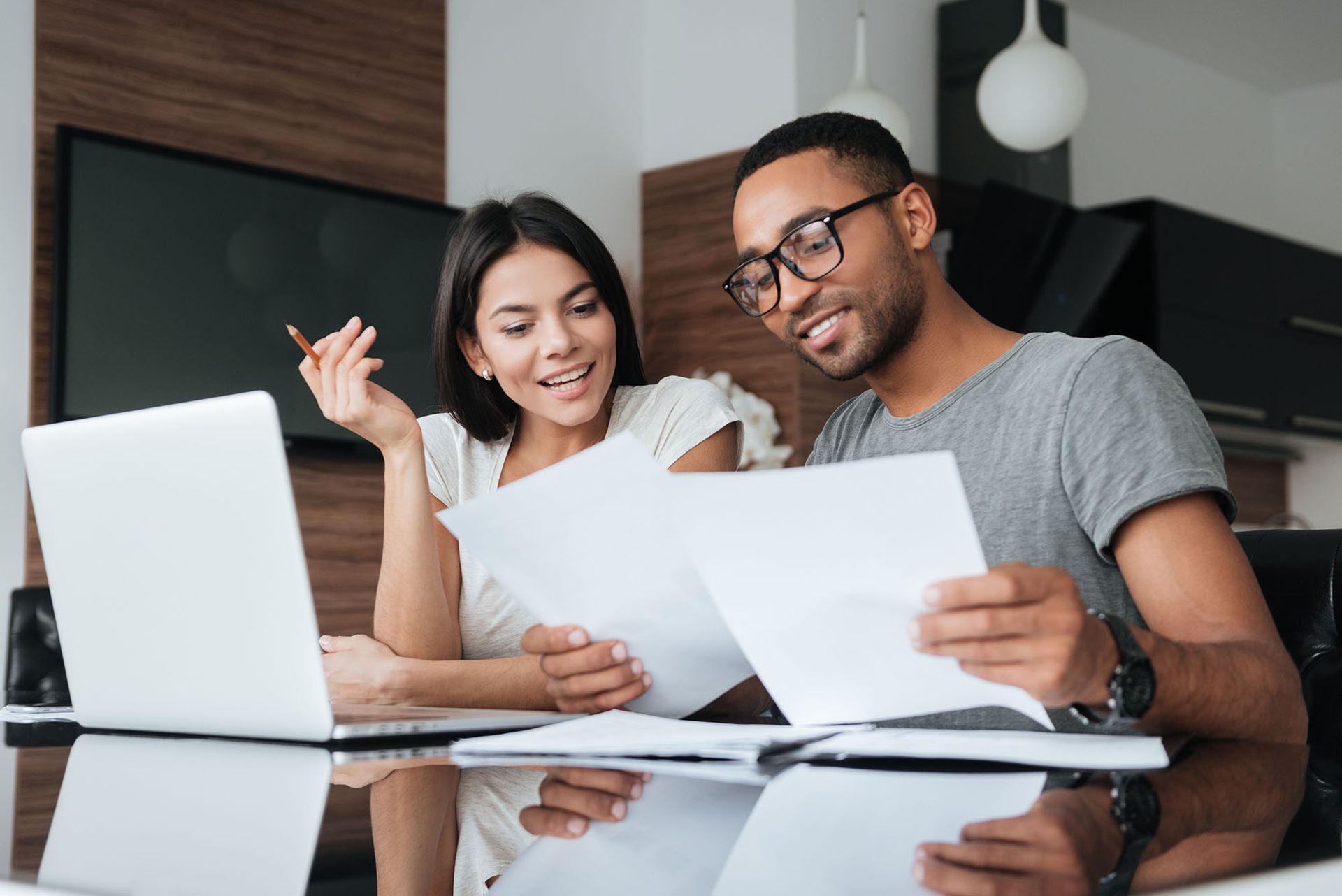 Couple reviews documents at a table with a laptop, smiling.