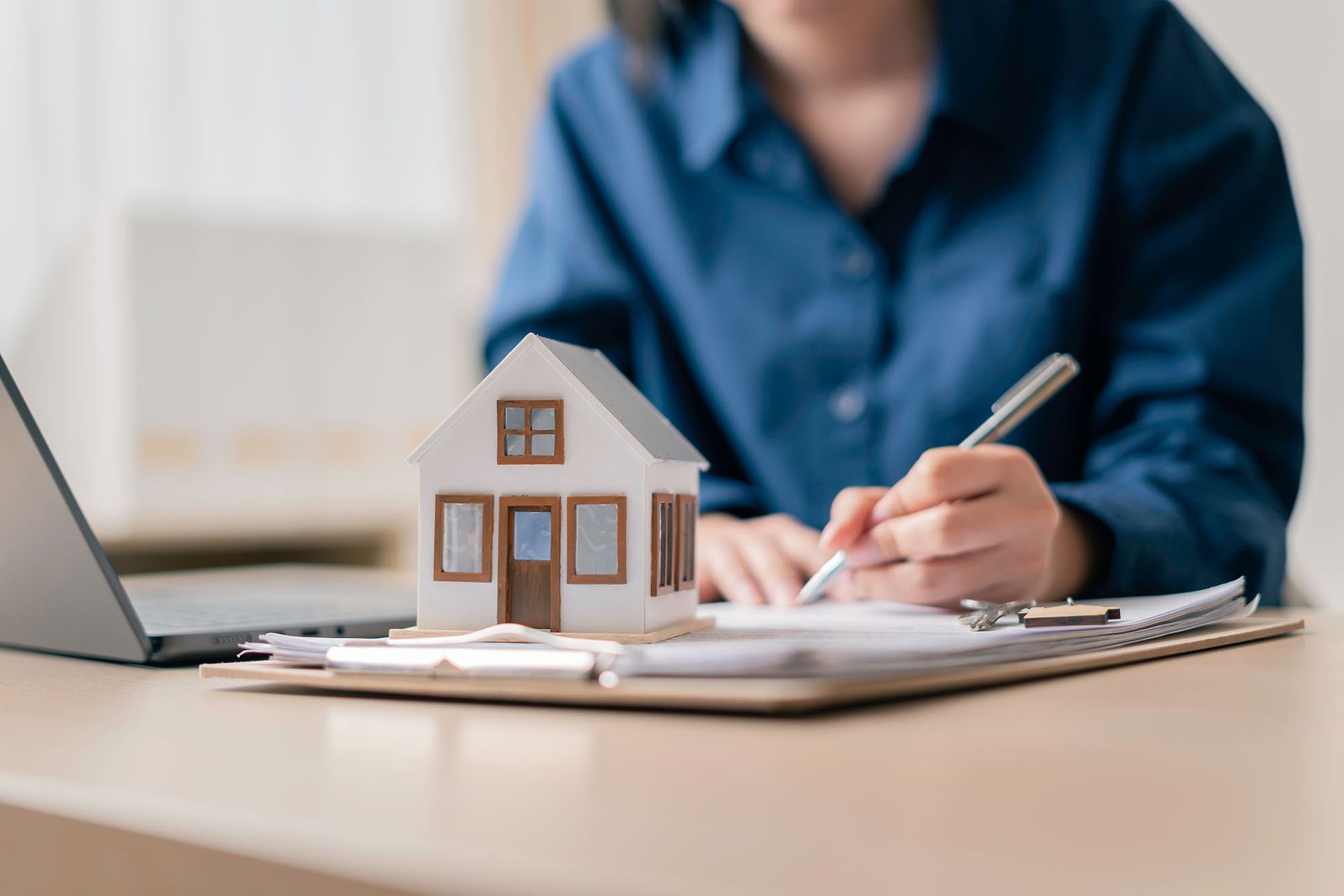 Miniature house on documents as person writes at desk.