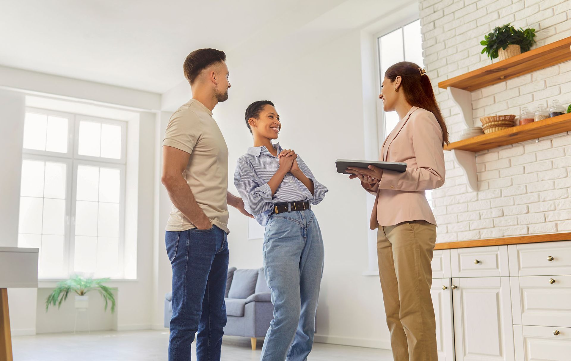 Couple and realtor discussing in an empty, bright kitchen. Woman holds a tablet.