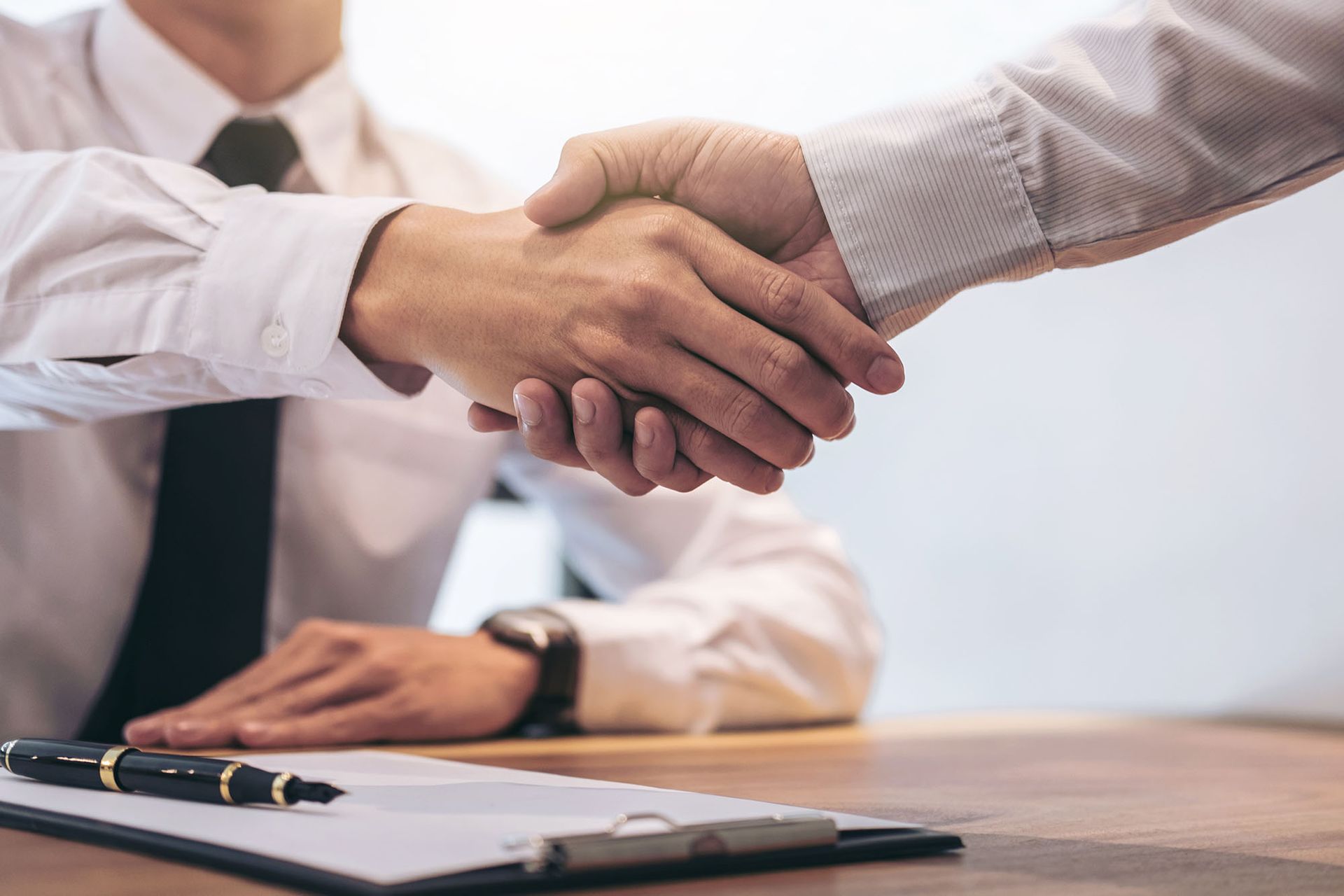 Two people in business attire shaking hands over a desk with a contract.