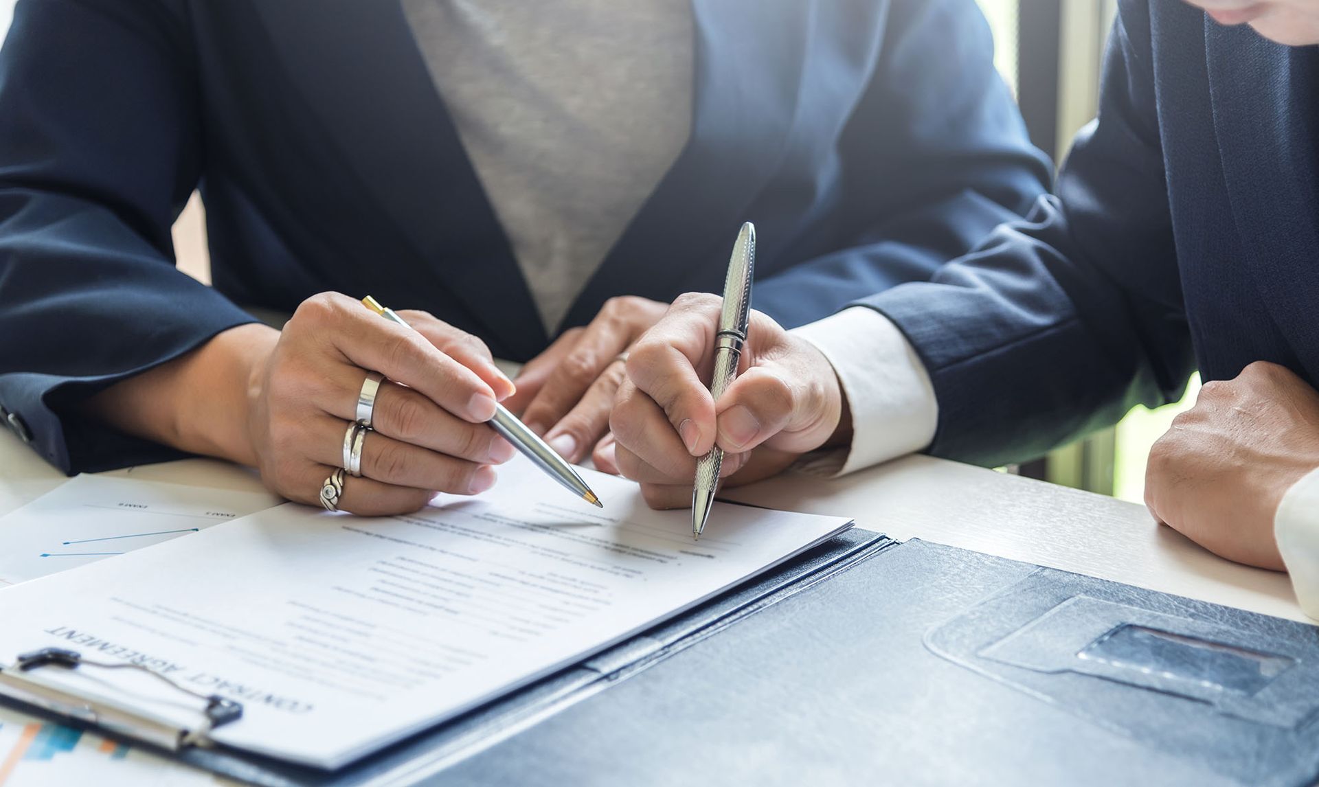 Two people in suits review a document at a table, pointing pens at the paper.