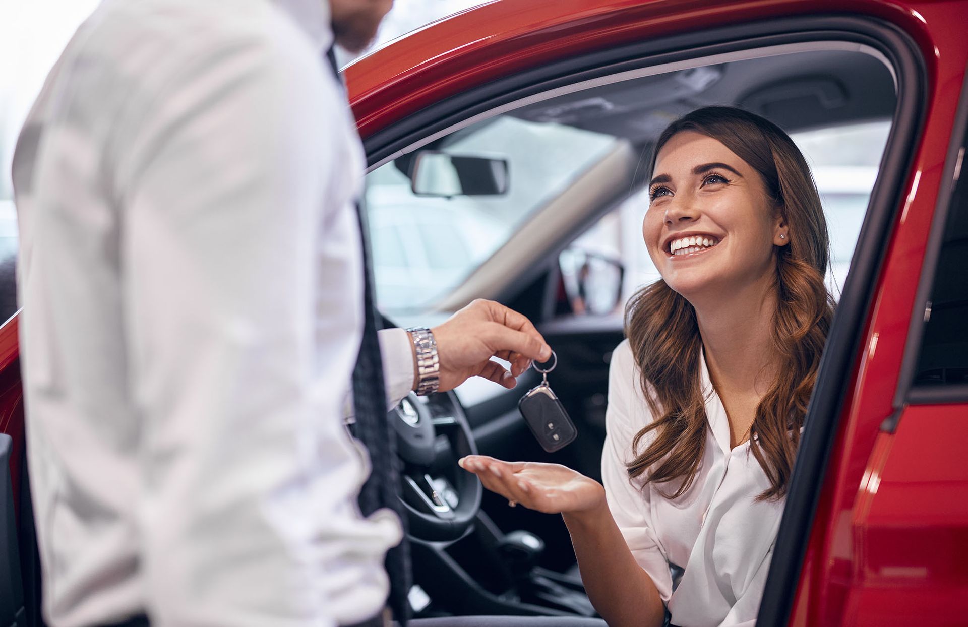 Car salesperson handing keys to a smiling person inside a red car.