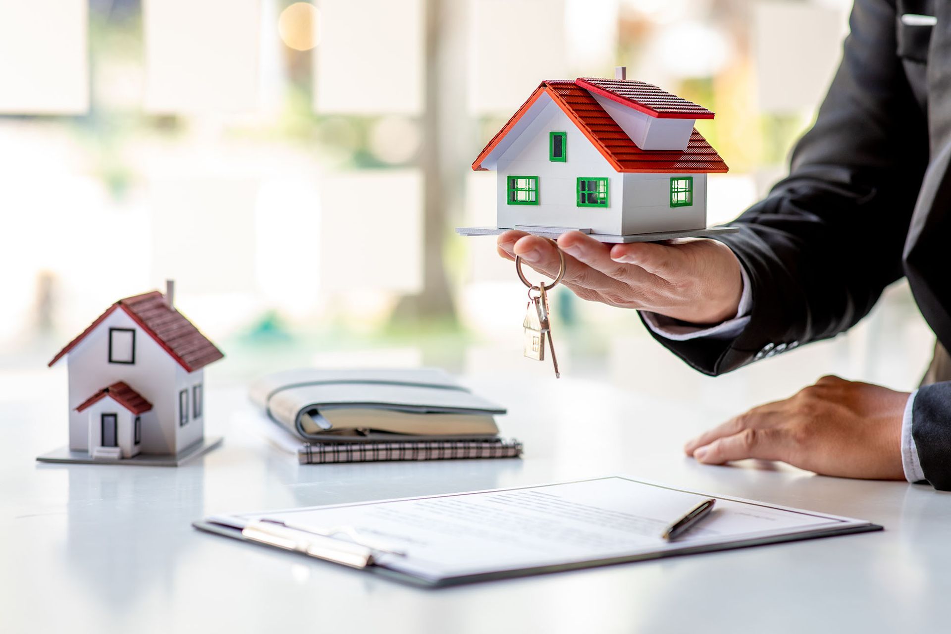 Real estate agent holding house model with keys, documents, and small house on a desk.