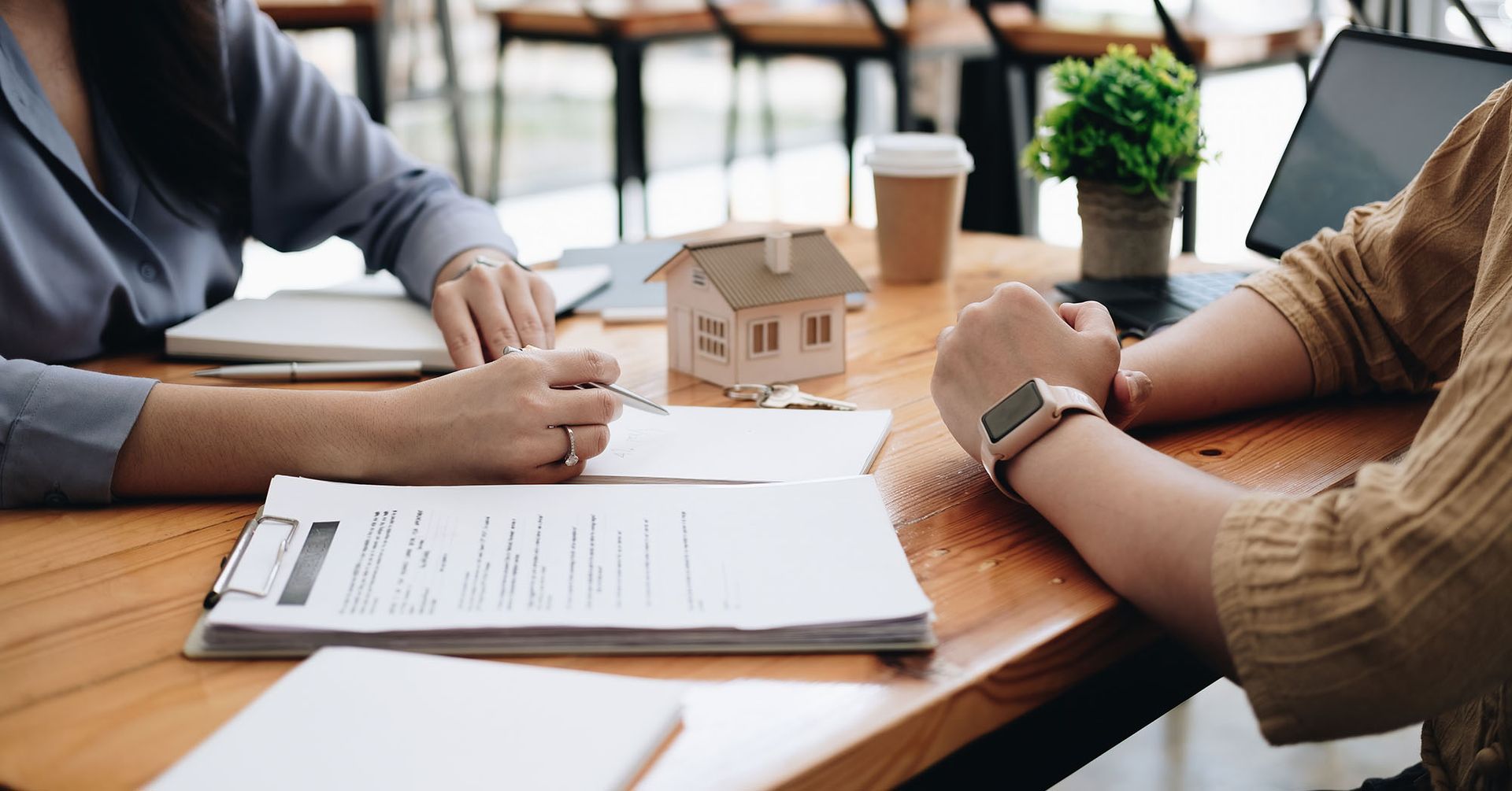 Person signing paperwork at a table with a house model, laptop, and coffee cup.