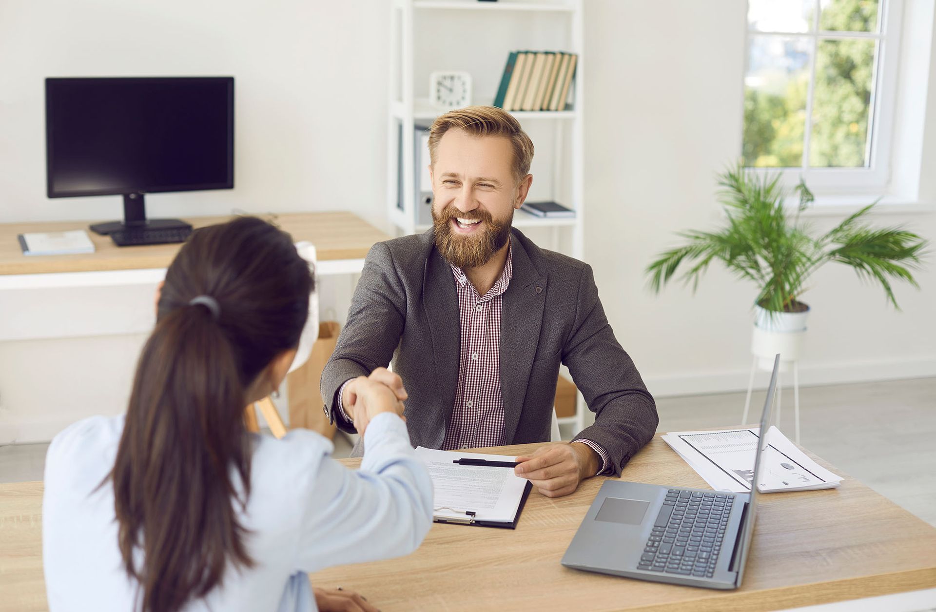 Woman and man shake hands across a desk. Man smiles; desk has laptop, papers, and a plant.