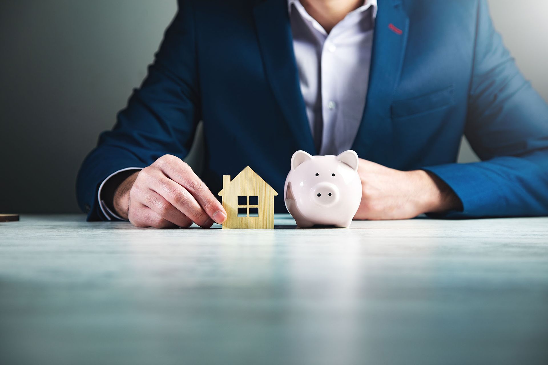 Person in suit holds a small house cutout and a piggy bank on a desk.