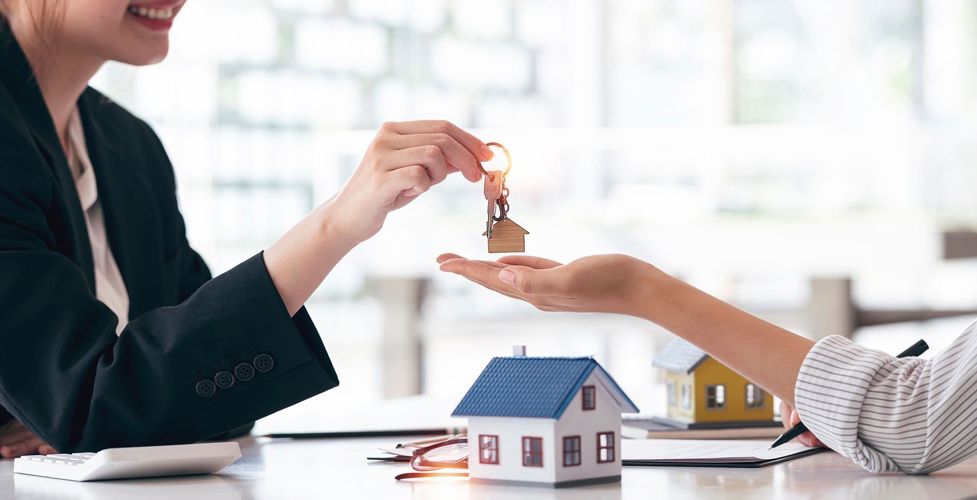 Person in blazer hands house keys to another person. Miniature house models sit on desk.