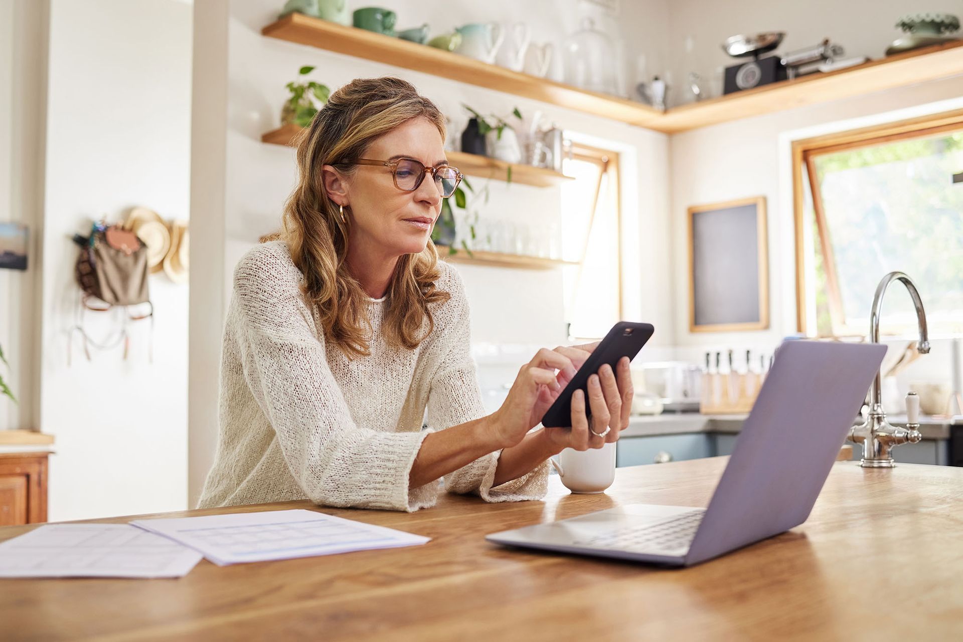 Woman in glasses, using phone and laptop at kitchen counter; papers nearby.