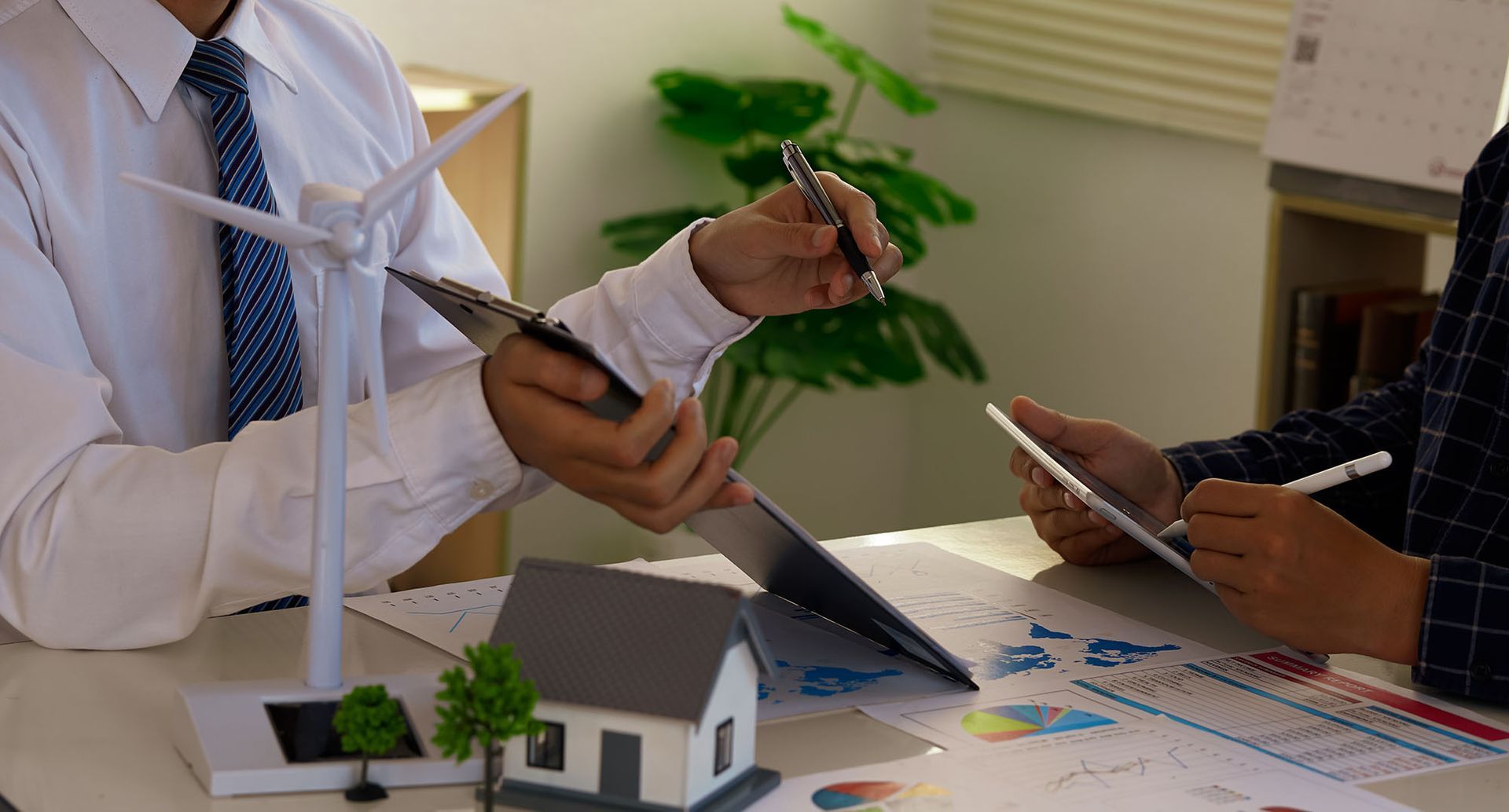 Two people review plans at a table with a wind turbine model, house model, and papers.