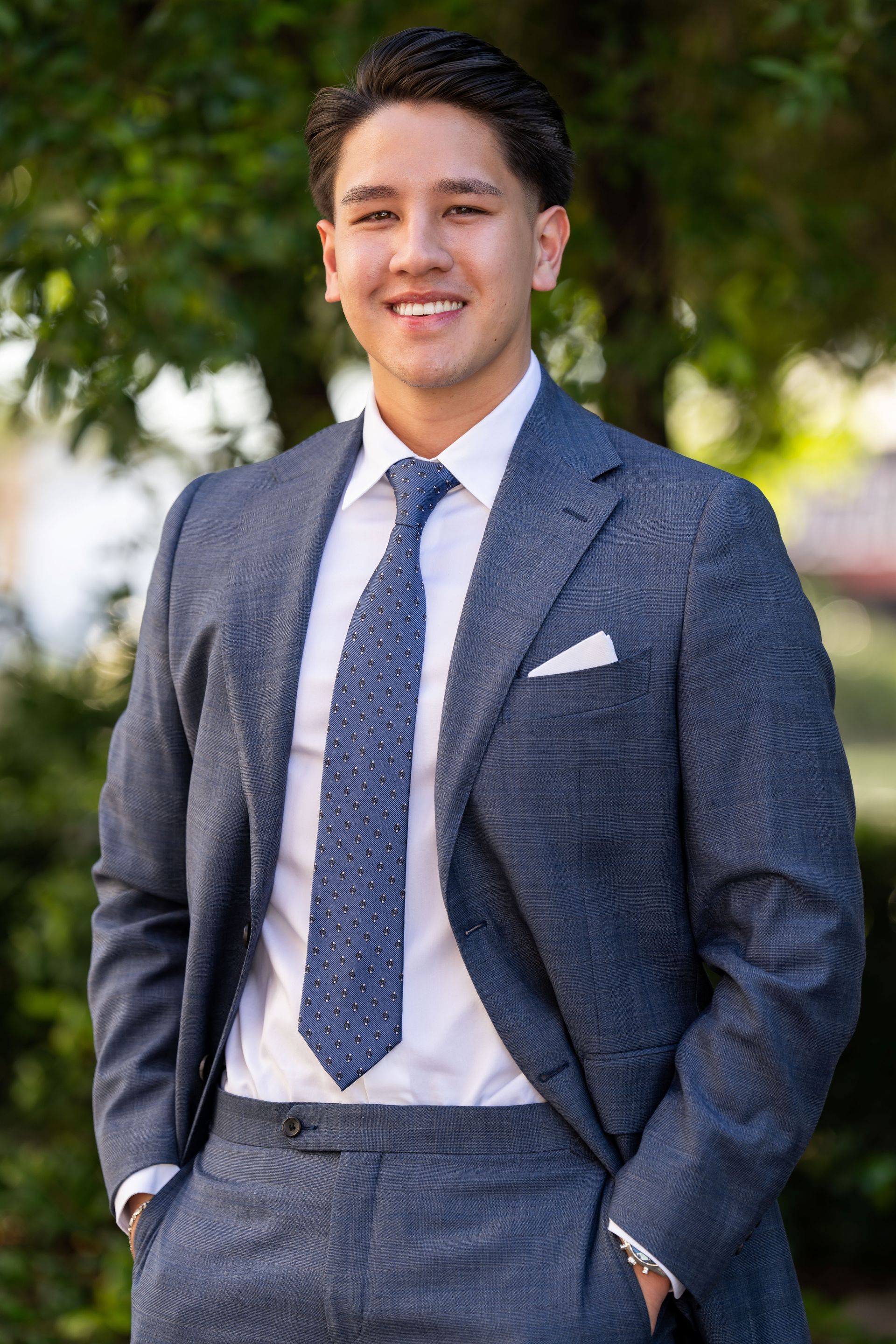 Man in navy suit and blue striped tie, smiles outdoors near foliage.