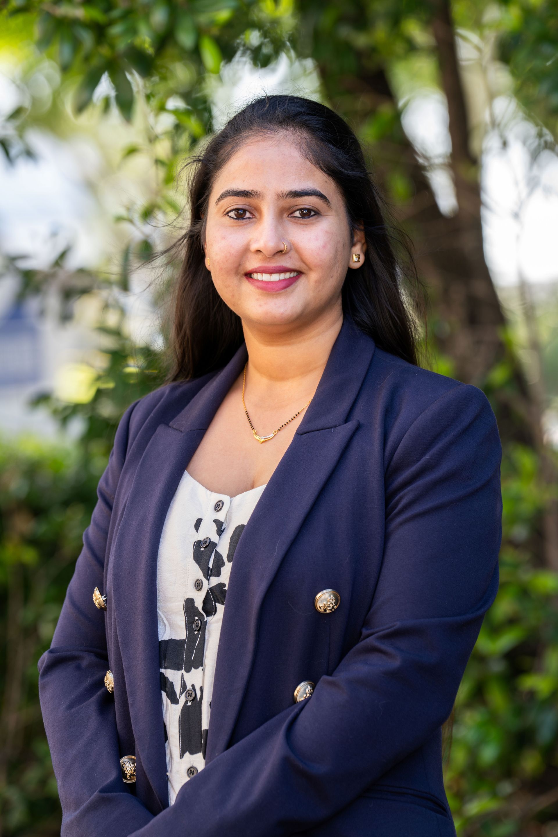 Man in a blue suit smiles at the camera, tie with stripes, green foliage background.