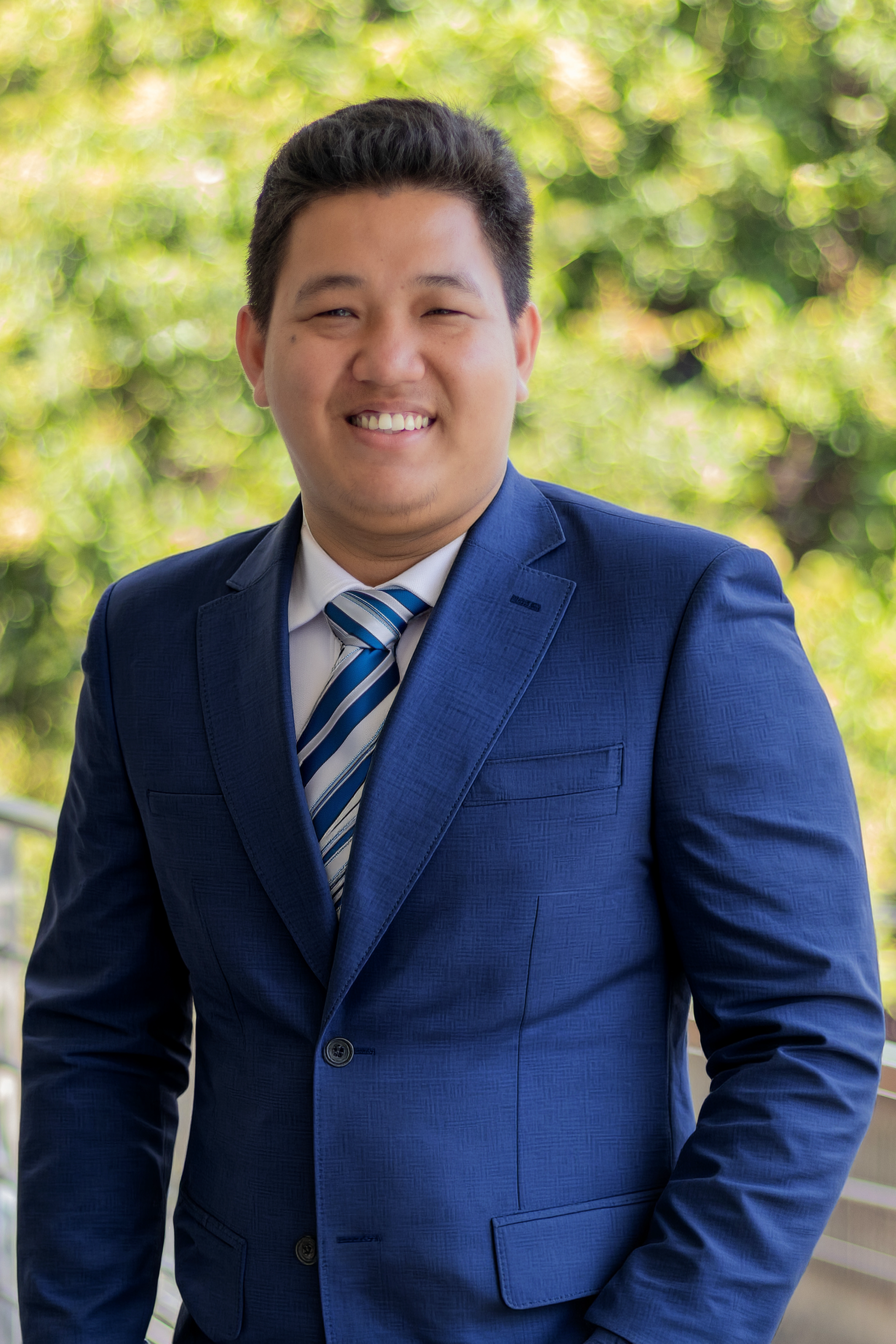 Man in navy suit and blue striped tie, smiles outdoors near foliage.