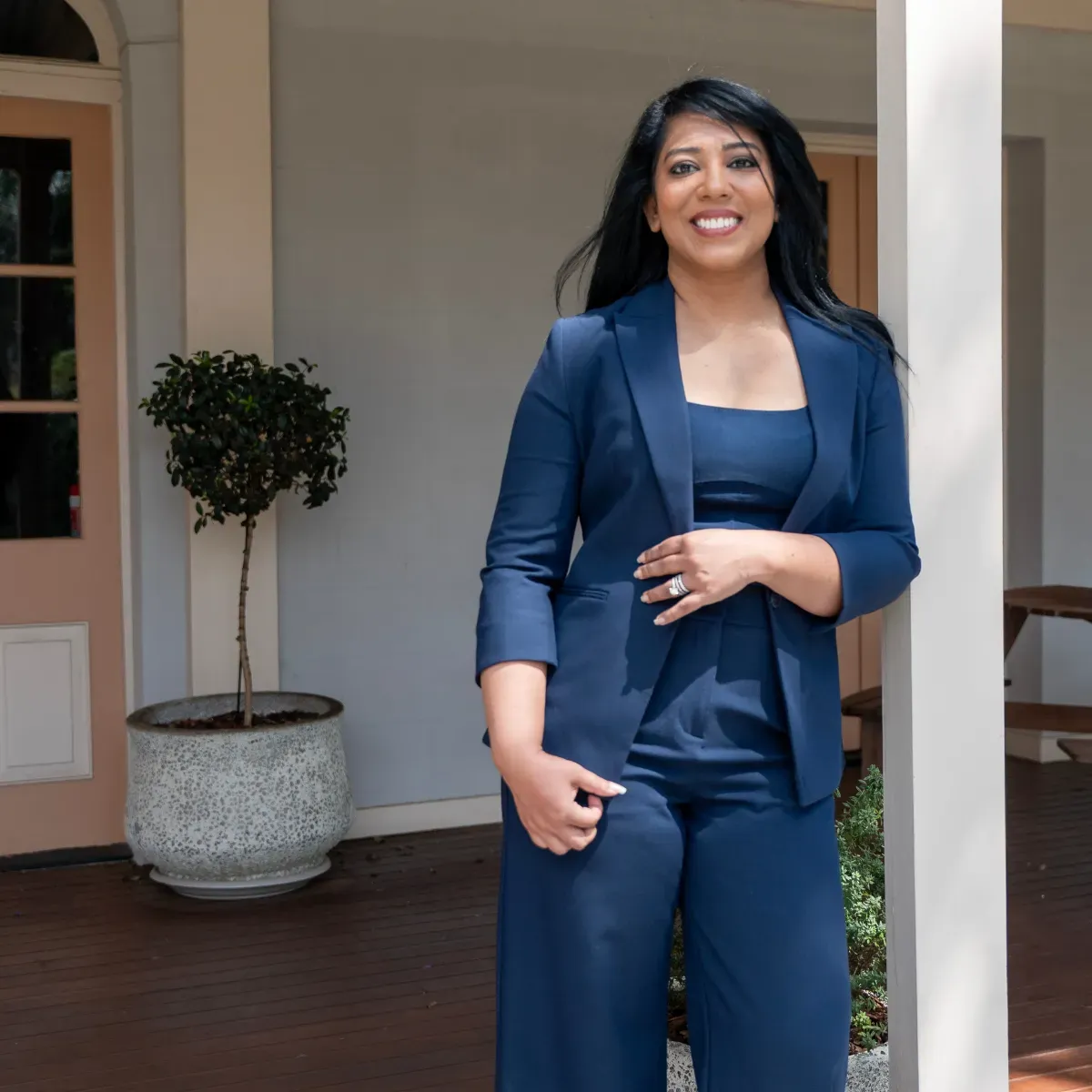 Woman in blue suit smiles, leans on a pillar outside a building with a pink door.