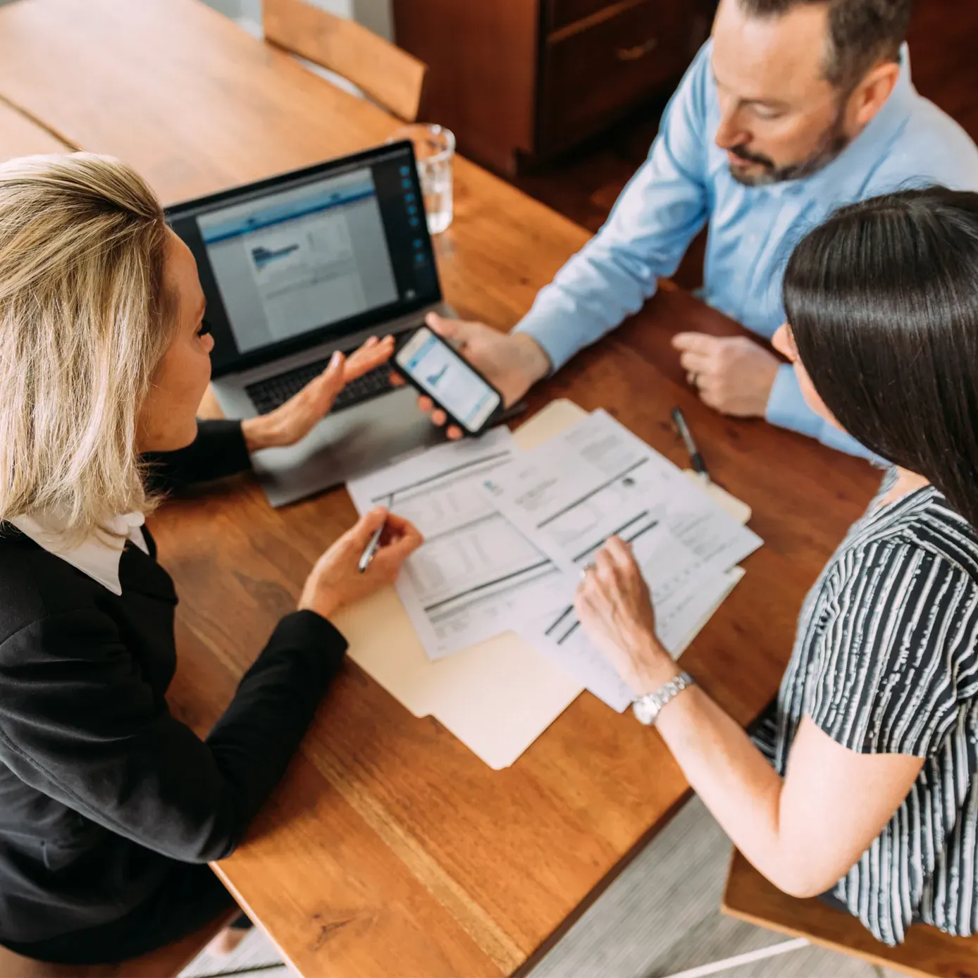 Three people reviewing documents around a wooden table. A laptop and phone are present.