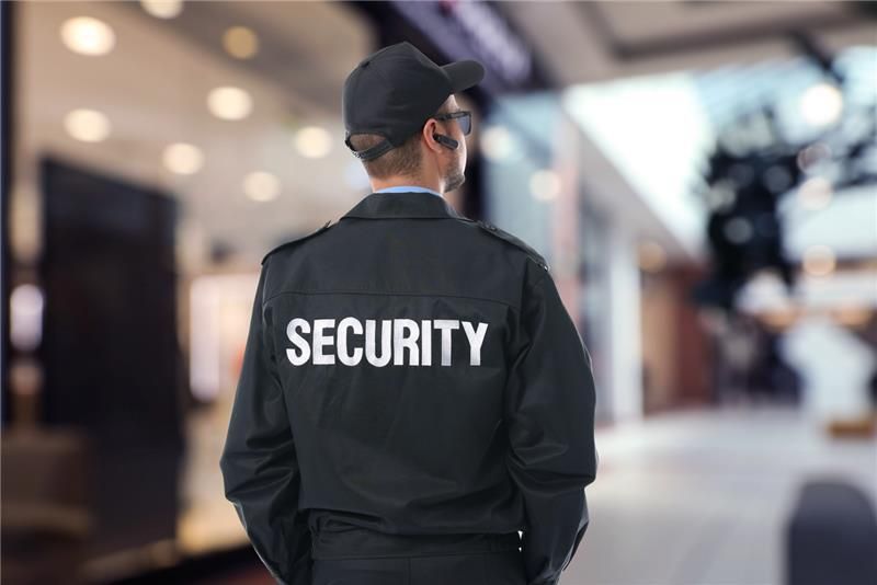 Security guard in black uniform stands in a mall, back to camera, looking ahead.